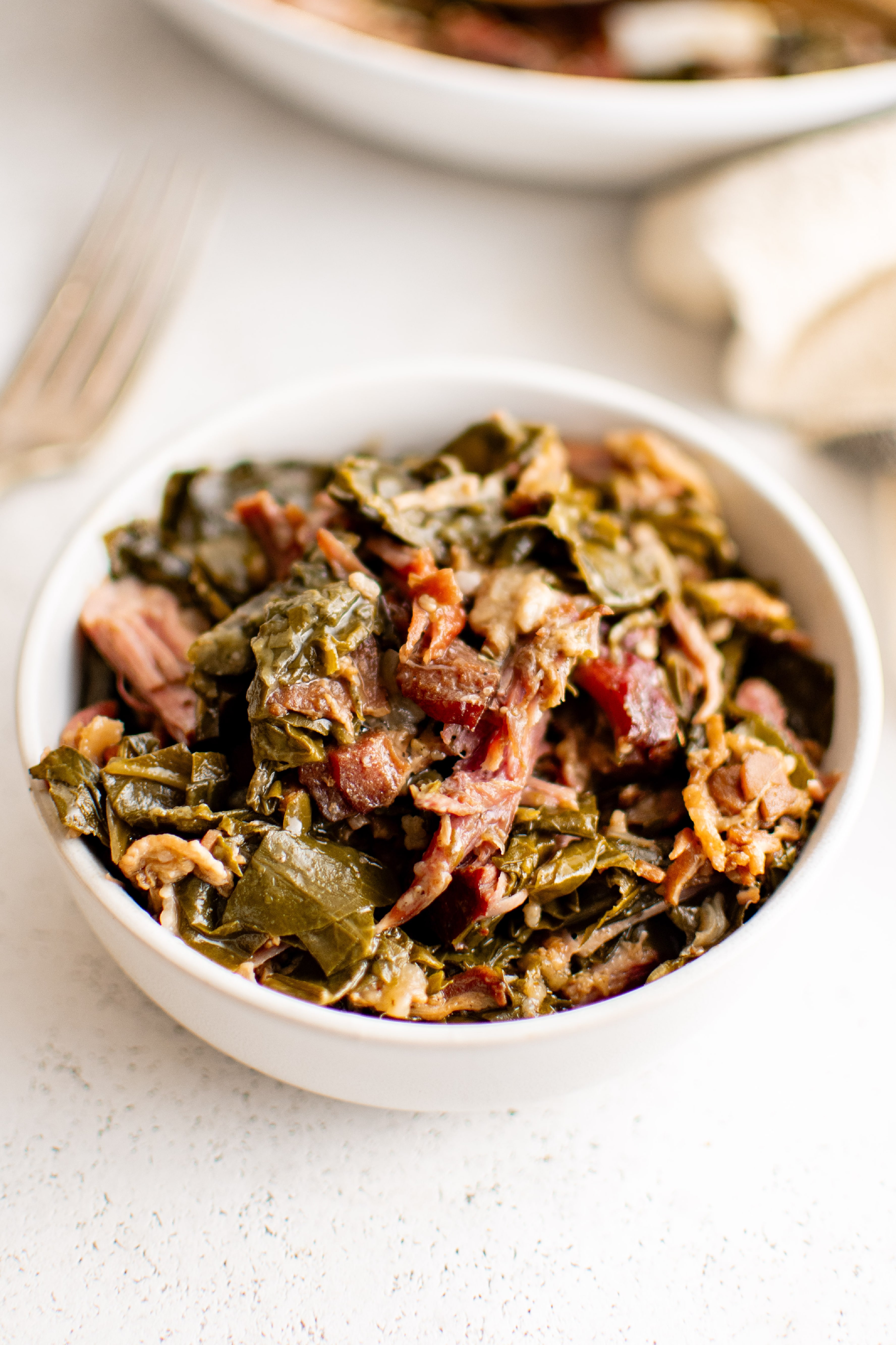 Small white serving bowl filled with cooked collard greens with shredded cooked ham hock and bacon.
