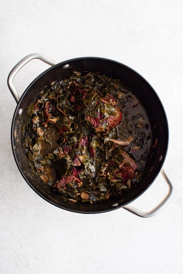 Large pot filled with cooked collard greens with bacon and three ham hocks.