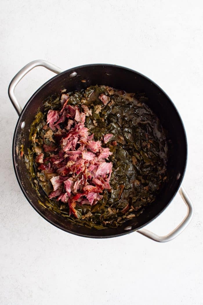 Shredded smoked ham hock meat added to a large pot filled with cooked collard greens.