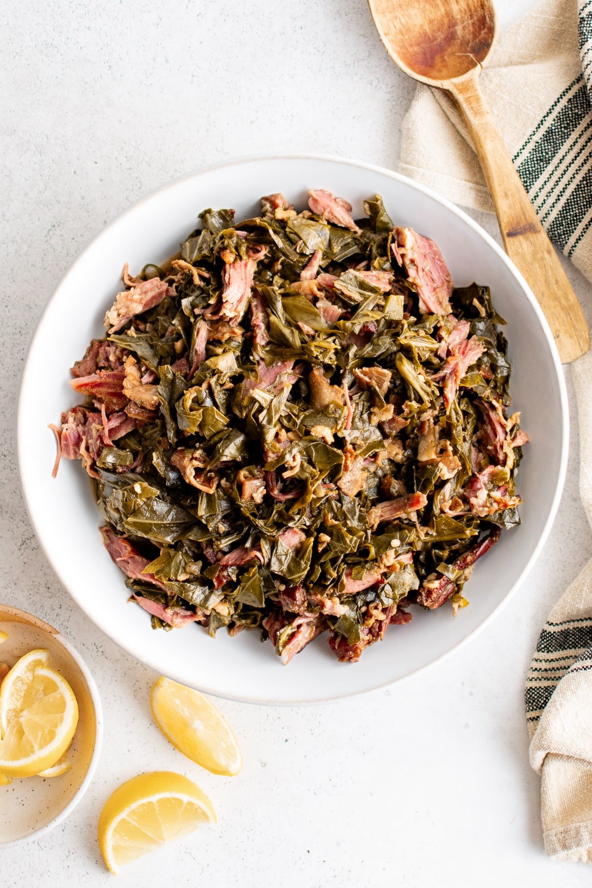 Overhead shot of collard greens in a white bowl with lemon wedges and a wooden spoon nearby