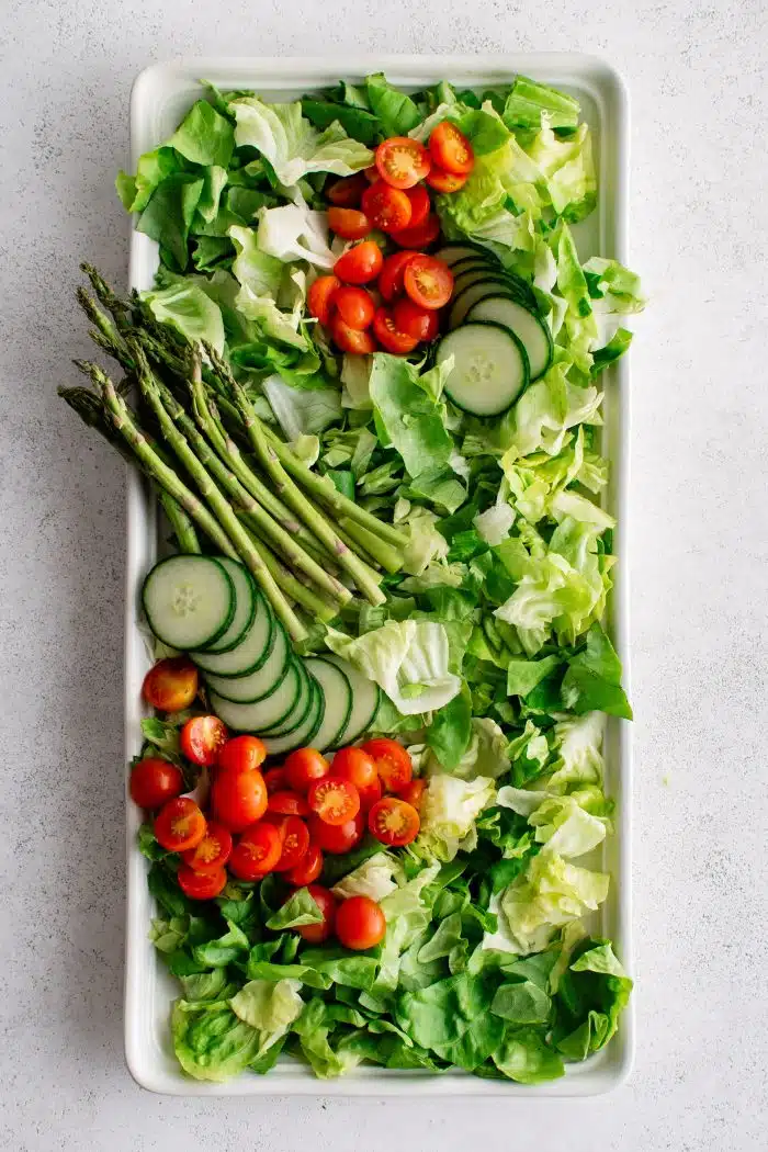 Large white rectangle salad platter plated with fresh butter lettuce and topped with sliced cucumber, asparagus, and cherry tomatoes.
