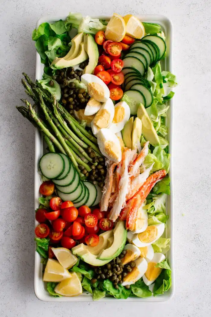 Large white rectangle salad platter plated with fresh butter lettuce and topped with sliced cucumber, asparagus, cherry tomatoes, sliced avocado, wedges of fresh lemons, and fresh cooked crab meat.