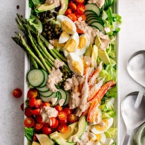 Large white rectangle salad platter plated beautifully with crab Louie salad made with a bed of butter lettuce topped with cherry tomatoes, hard-boiled eggs, avocado, sliced cucumbers, lemon slices, asparagus, lump crab meat and drizzled with homemade dressing.