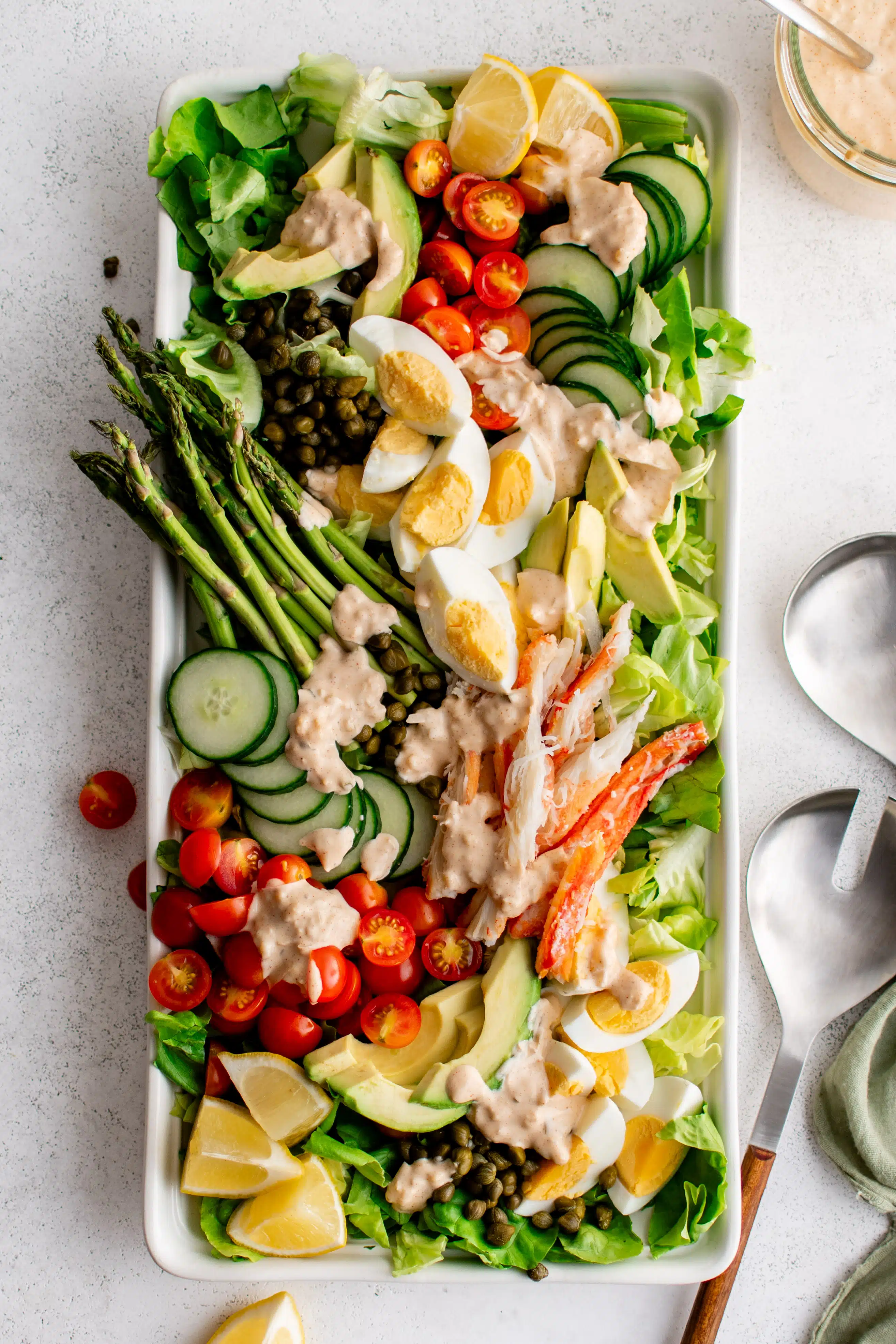 Large white rectangle salad platter plated beautifully with crab Louie salad made with a bed of butter lettuce topped with cherry tomatoes, hard-boiled eggs, avocado, sliced cucumbers, lemon slices, asparagus, lump crab meat and drizzled with homemade dressing.