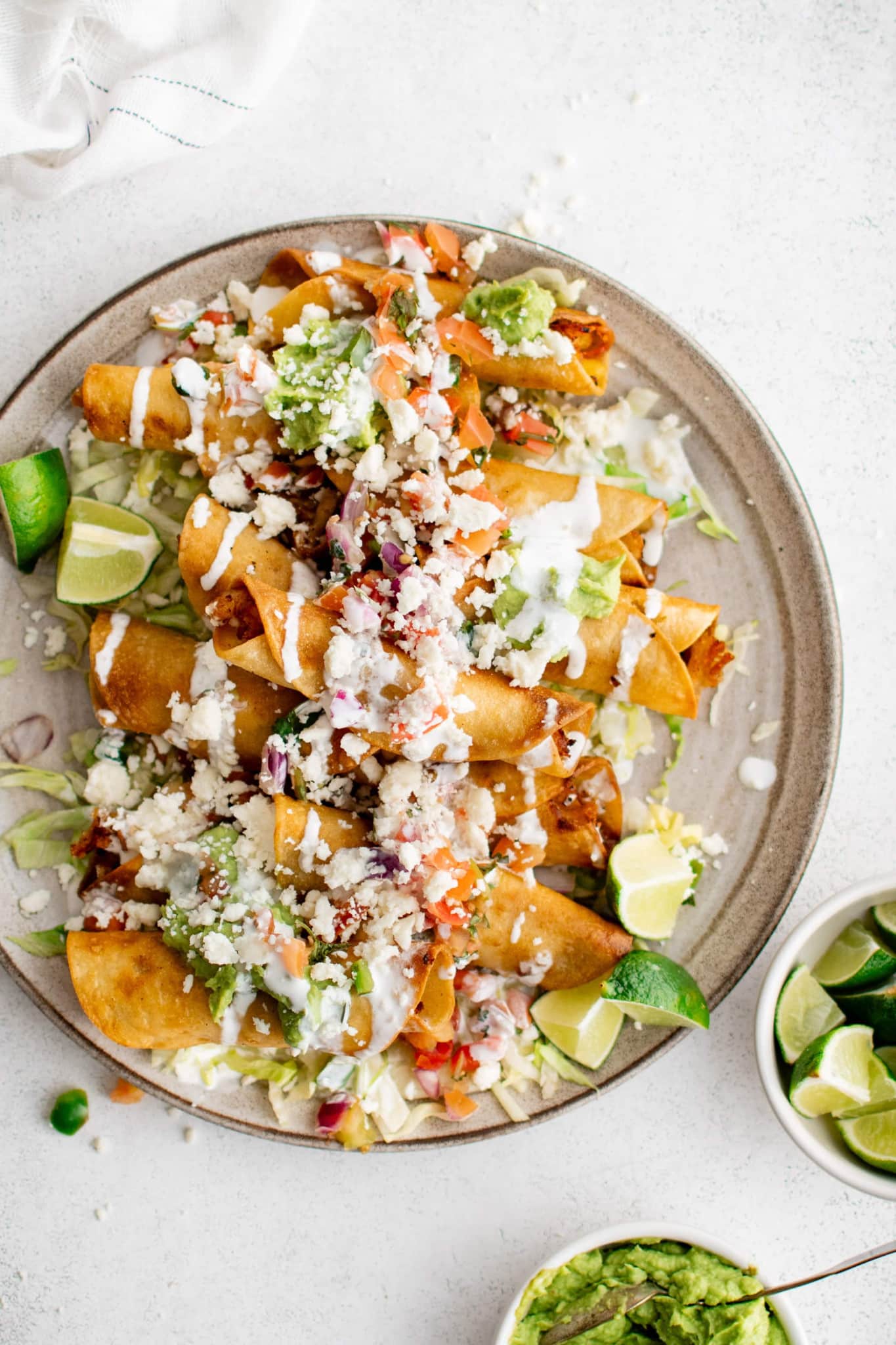 Plate filled with deep-fried chicken and potato flautas with shredded iceberg lettuce, crumbled cotija cheese, tomatoes, lime wedges, and guacamole.