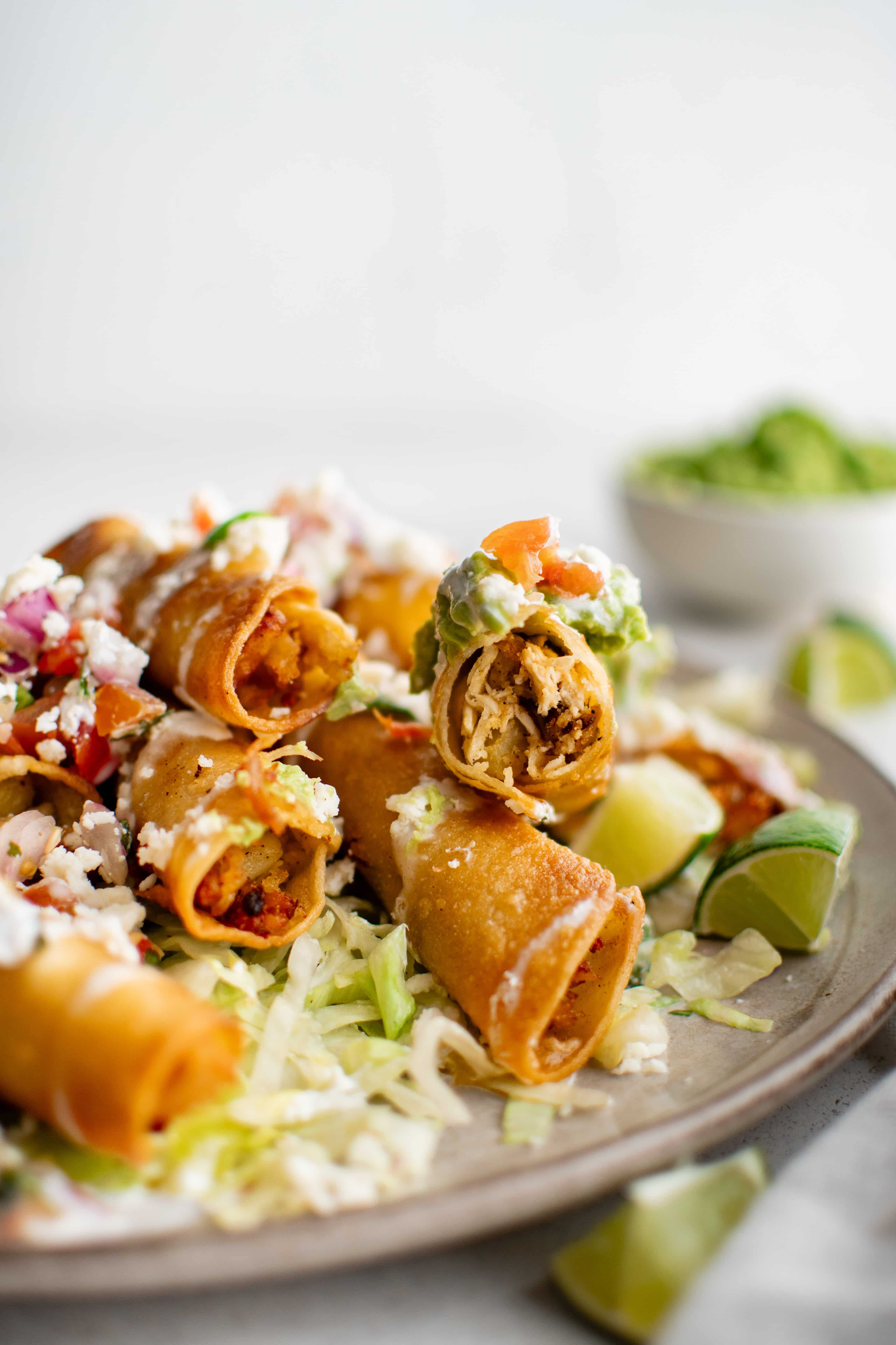 Plate filled with deep-fried chicken and potato flautas de pollo with shredded iceberg lettuce, crumbled cotija cheese, tomatoes, lime wedges, and guacamole.