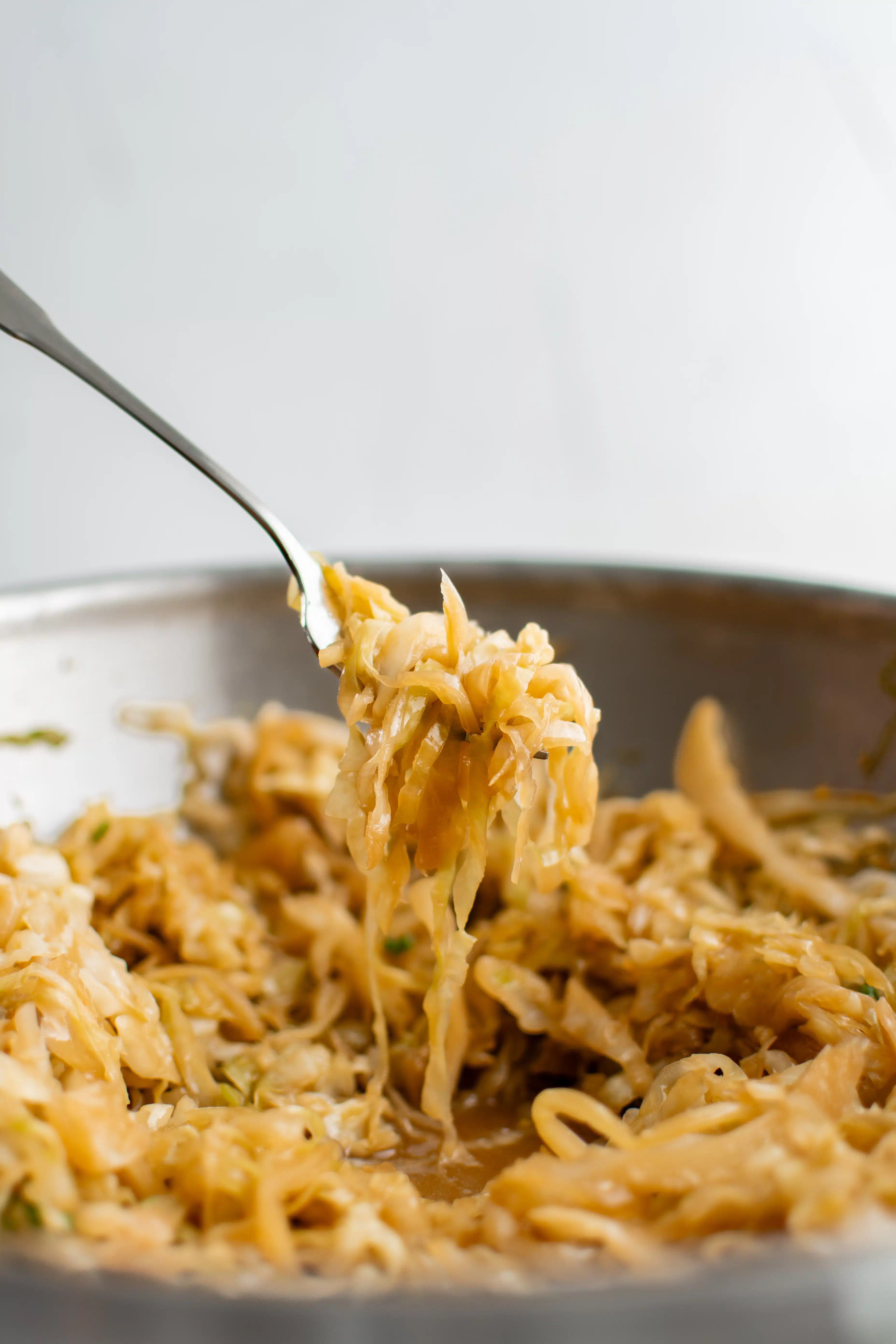 Fork-full of soft stir-fried cabbage hovering above a large wok filled with cooked cabbage stir-fry.