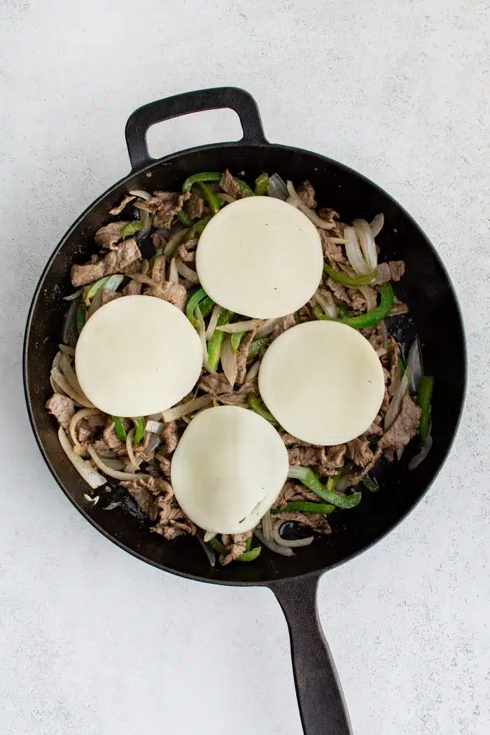 Cooked onions, green bell peppers, and sliced sirloin steak in a large cast iron skillet topped with four slices of mild provolone cheese.