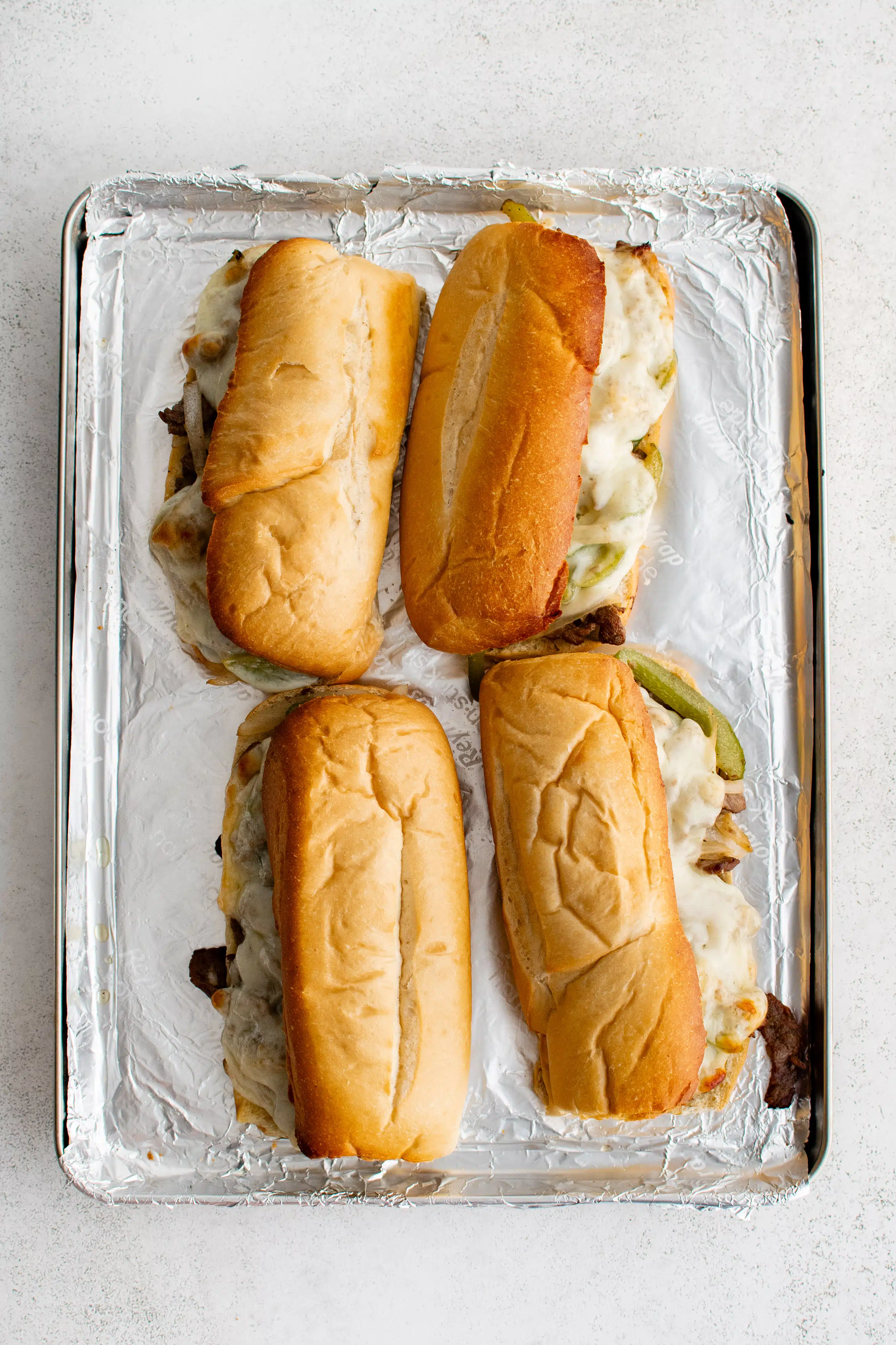 Four Philly cheesesteak sandwiches on a large baking sheet lined with foil.