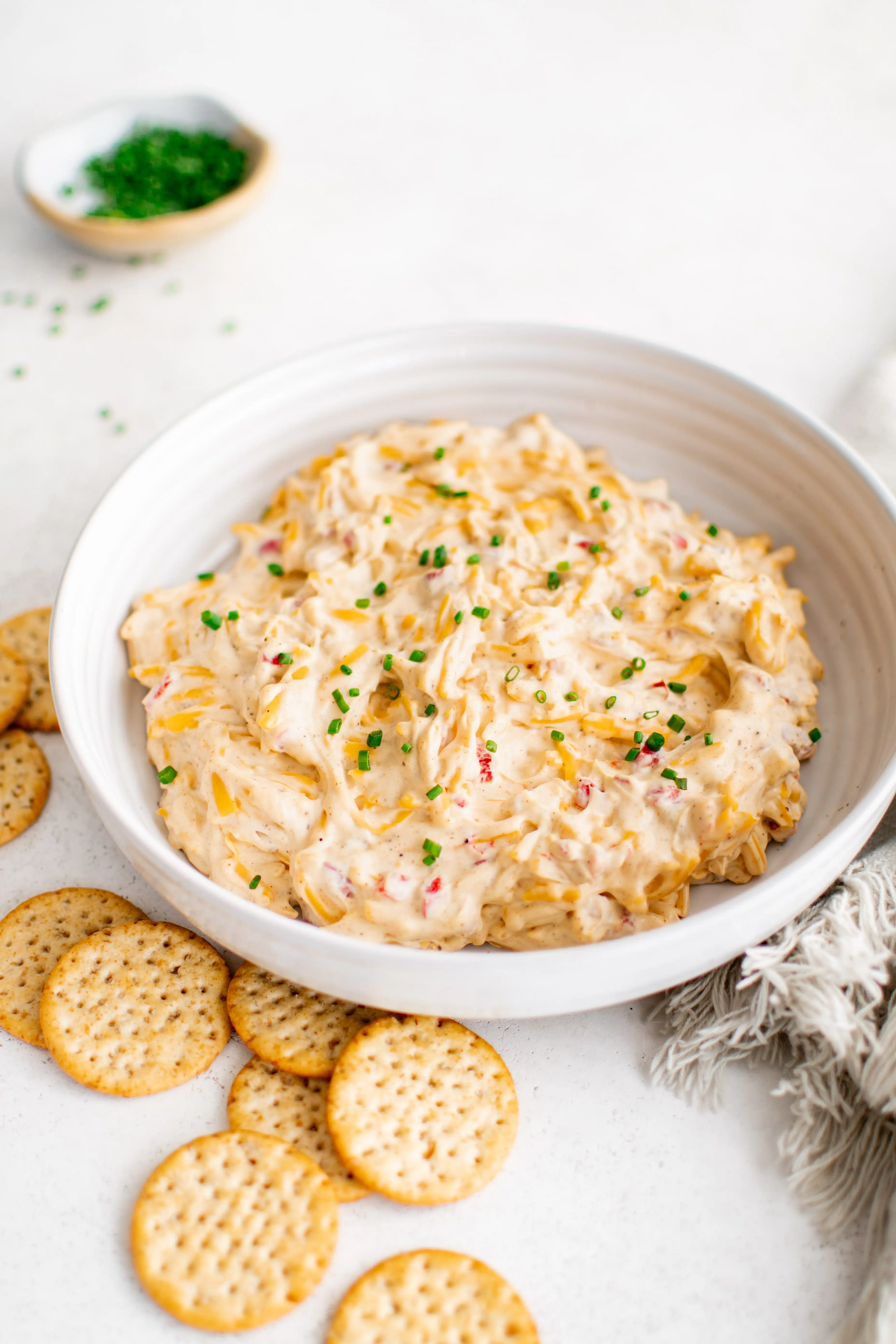 Large shallow mixing bowl filled with pimento cheese dip made with cream cheese, mayonnaise, cheddar cheese, and diced pimentos and garnished with freshly chopped chives and served with crackers on the side.