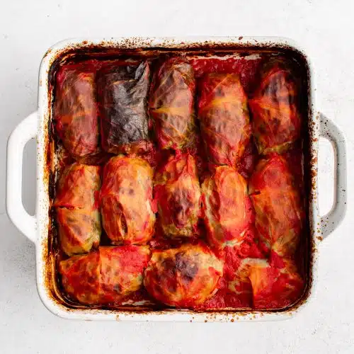 White baking dish filled with cabbage rolls stuffed with ground beef, pork, and rice and covered in tomato sauce.