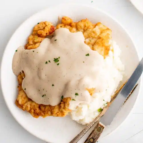 White plate with a scoop of mashed potatoes topped with a golden piece of chicken fried chicken smothered in white gravy.