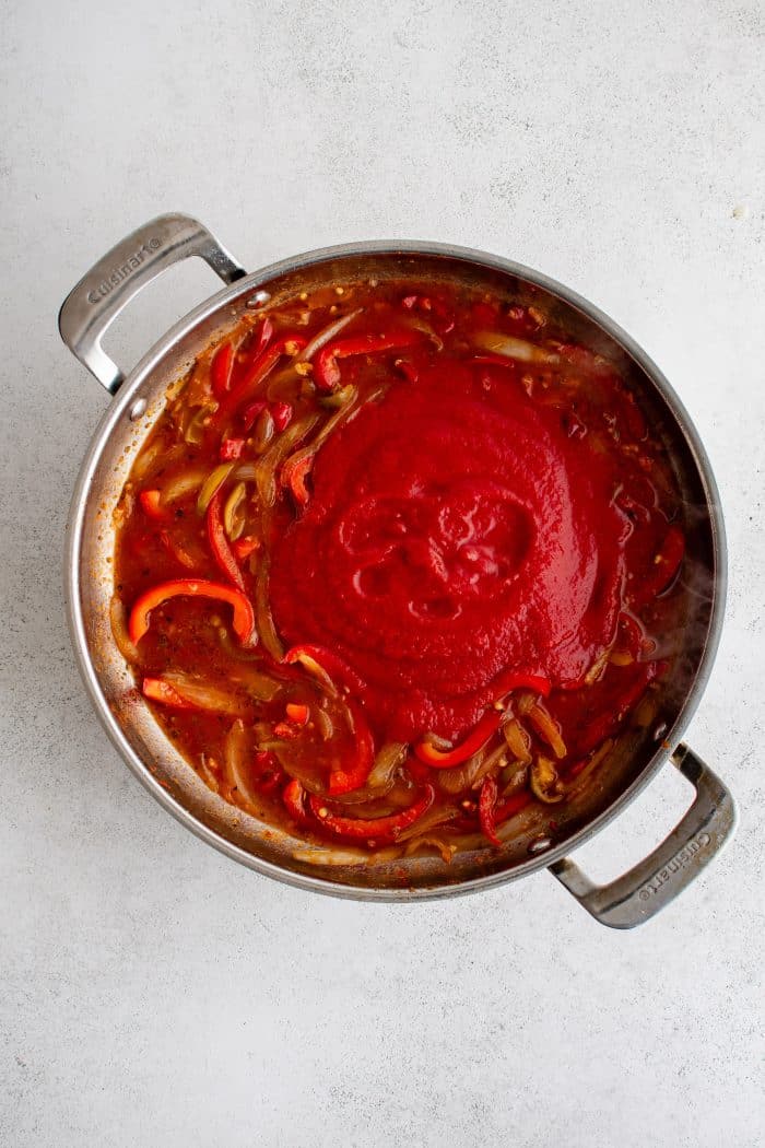 Tomato sauce added to a large stainless steel pan filled with sauteed bell peppers, onions, hot cherry peppers, and tomato paste.