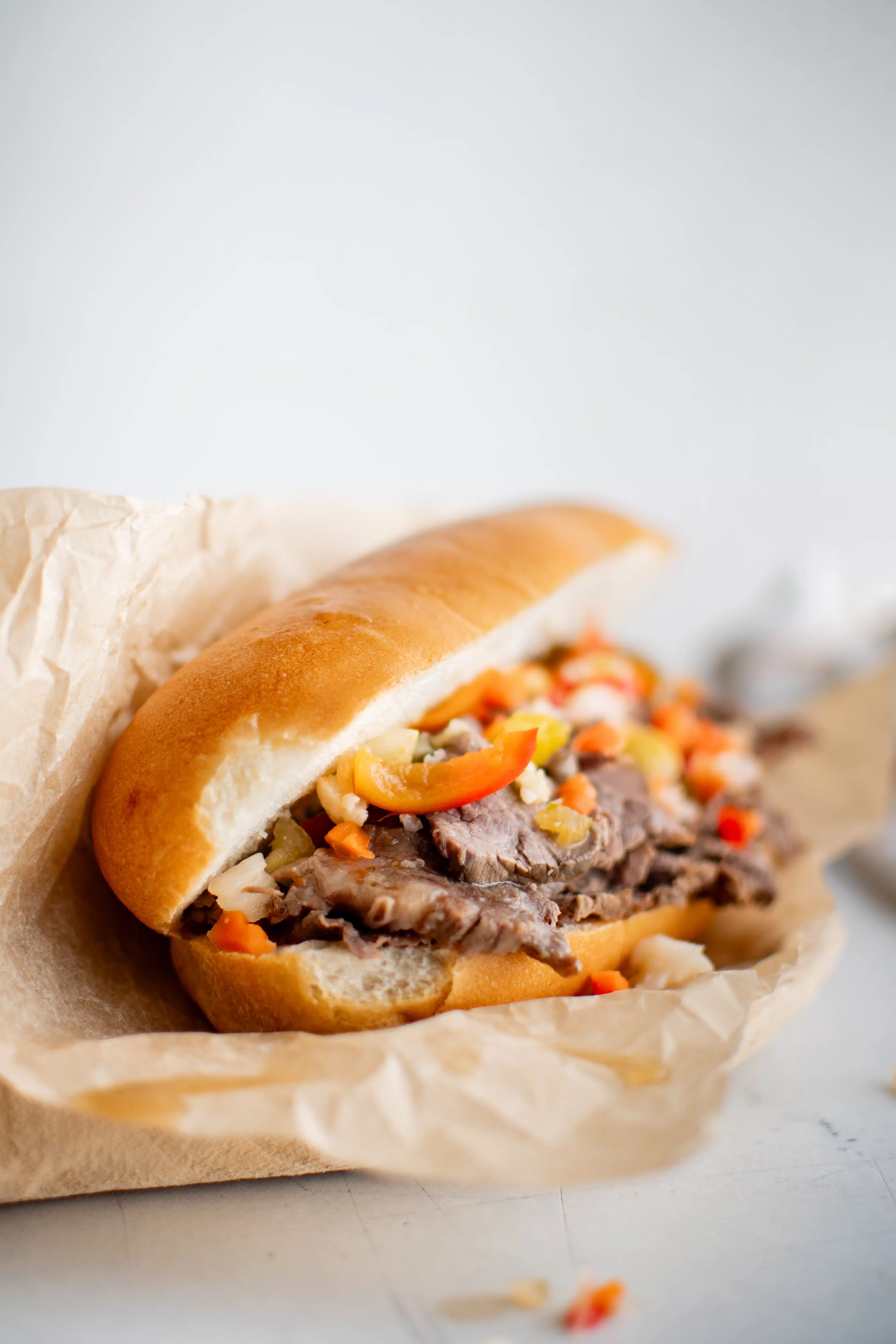 Italian roll topped with thinly sliced beef topped with chopped Giardiniera and hot cherry peppers served on a white plate lined with parchment paper.