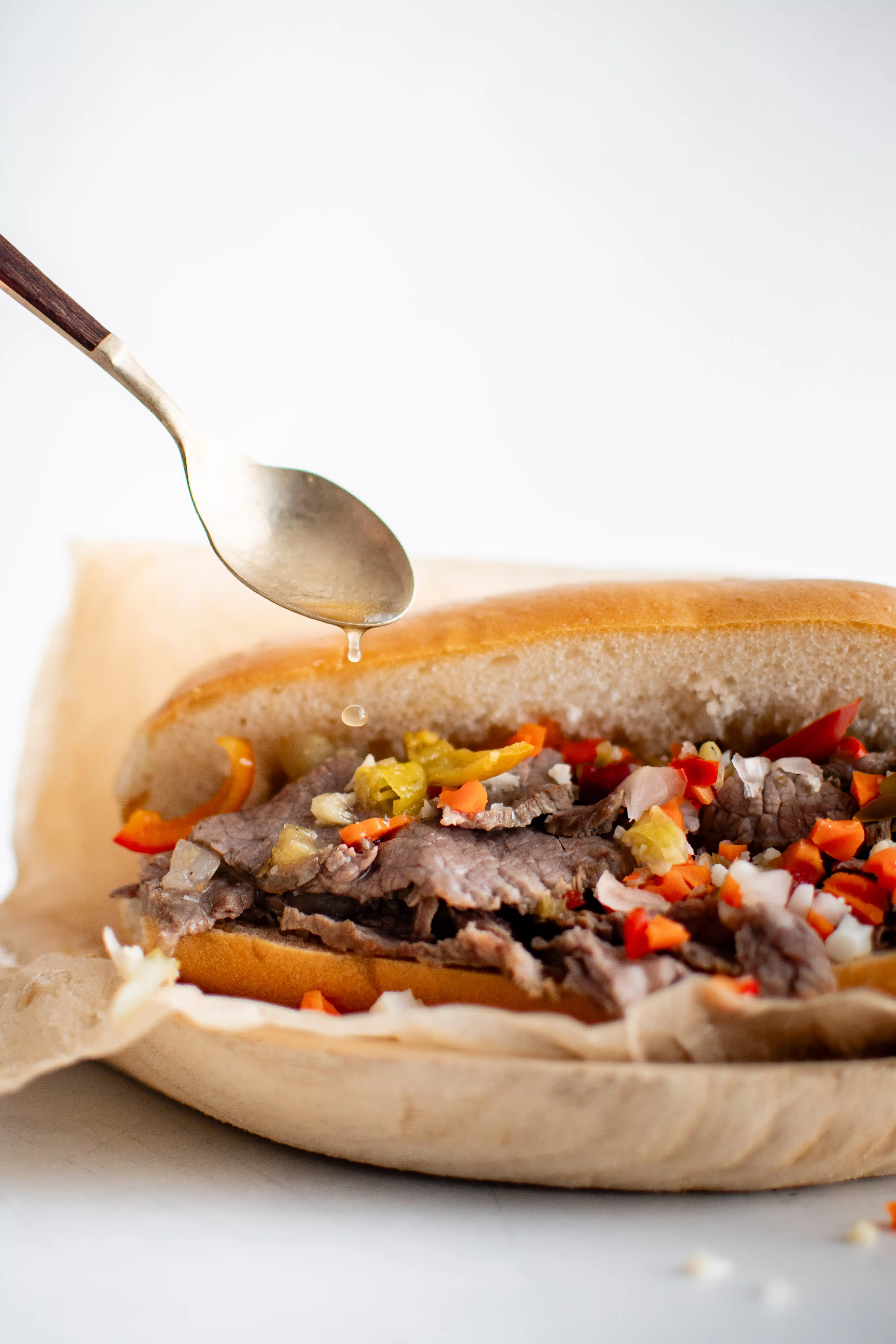 Drizzling au jus over an Italian roll topped with thinly sliced beef topped with chopped Giardiniera and hot cherry peppers served on a white plate lined with parchment paper.