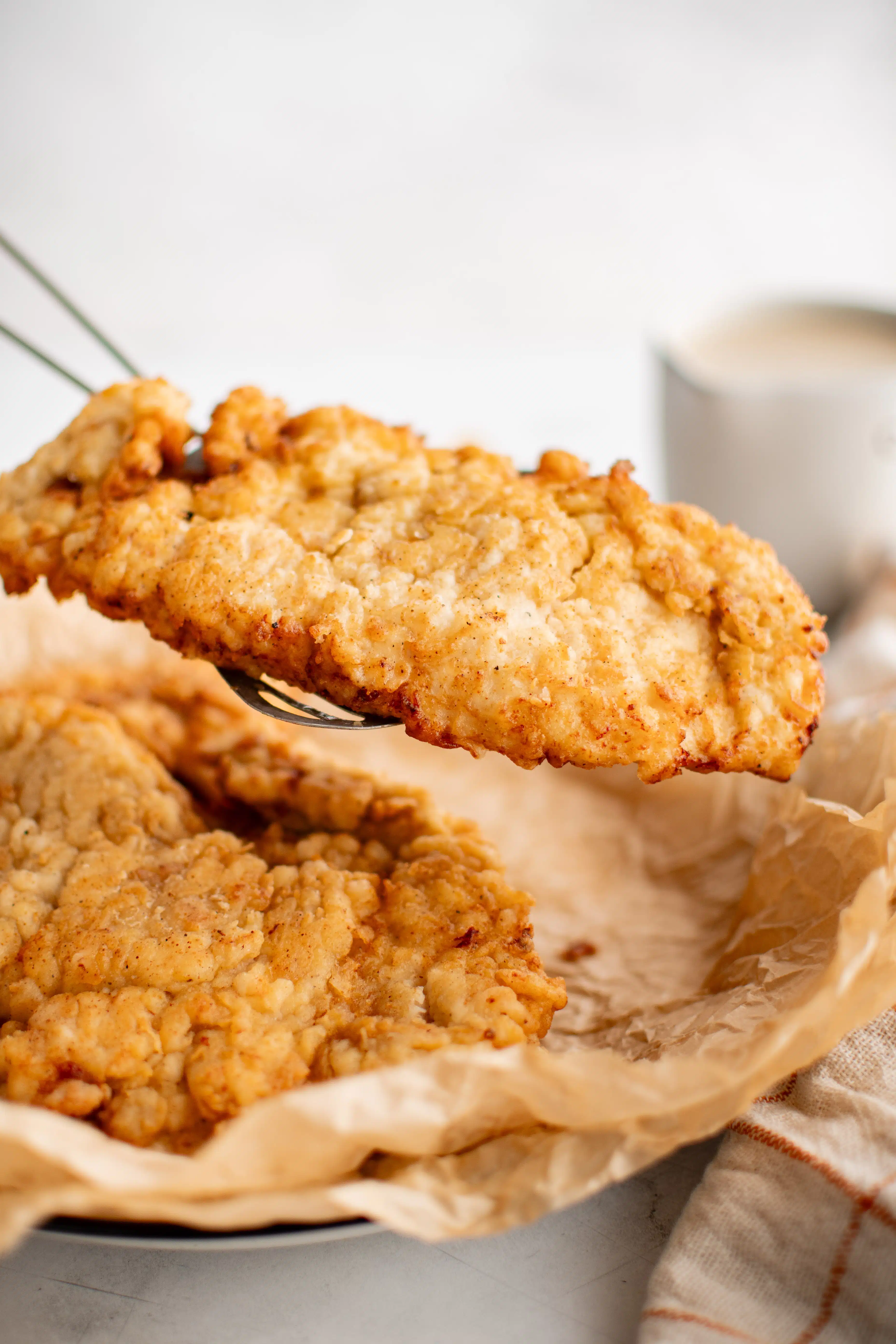 Metal tongs holding a breaded and fried piece of chicken fried chicken from a plate lined with brown wax paper.