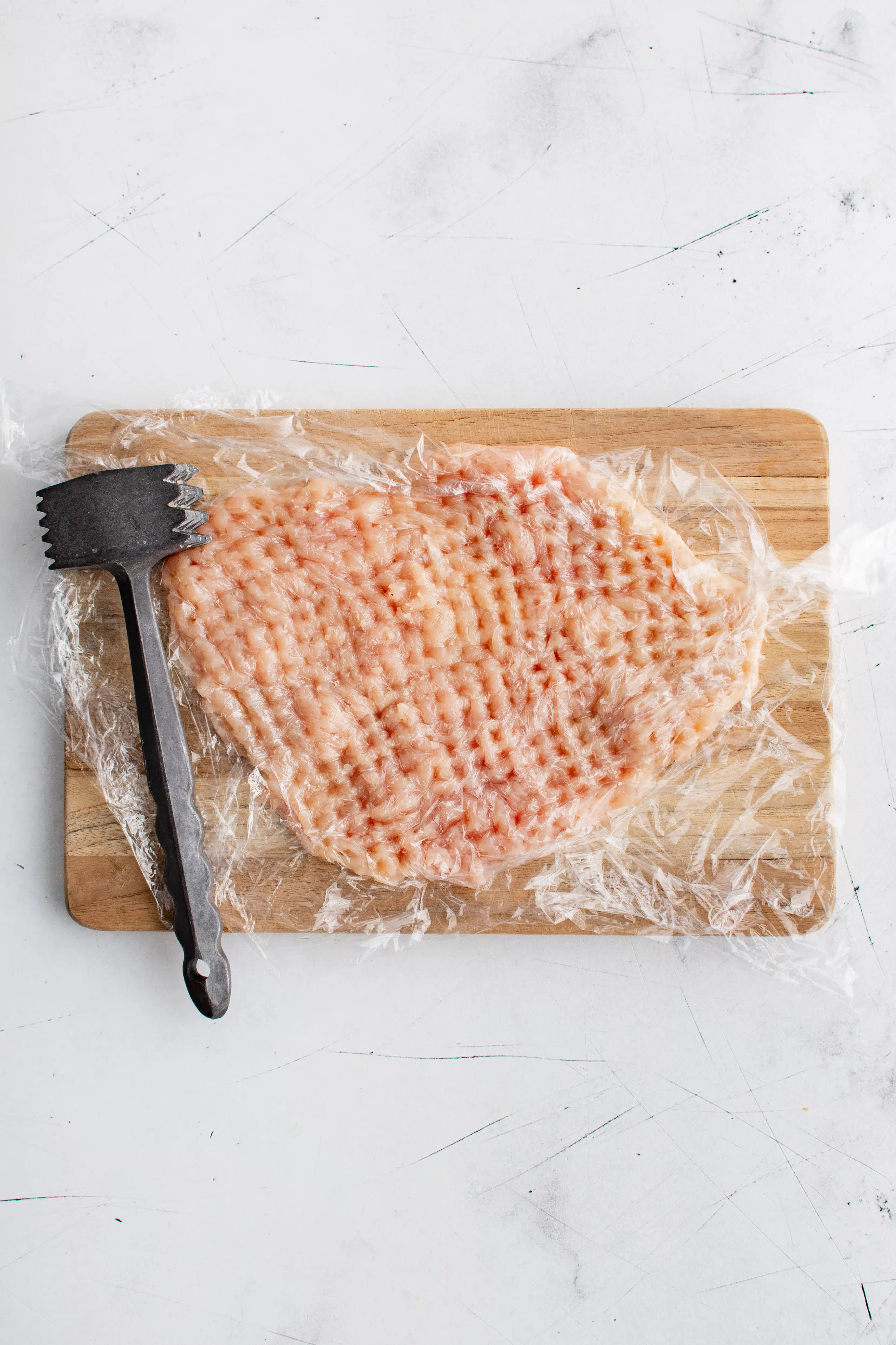 Large cutting board with a chicken breast under plastic wrap and pounded thin with a meat mallet.