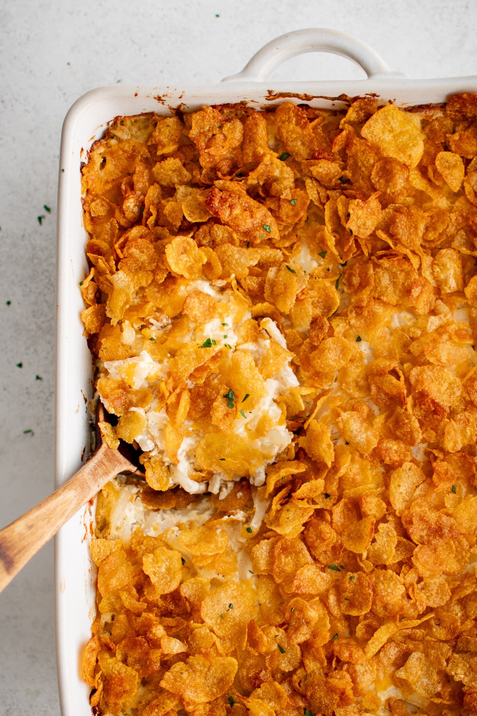 Wooden serving spoon in a large white casserole dish filled with gooey, cheesy baked funeral potatoes topped with crispy cornflakes.