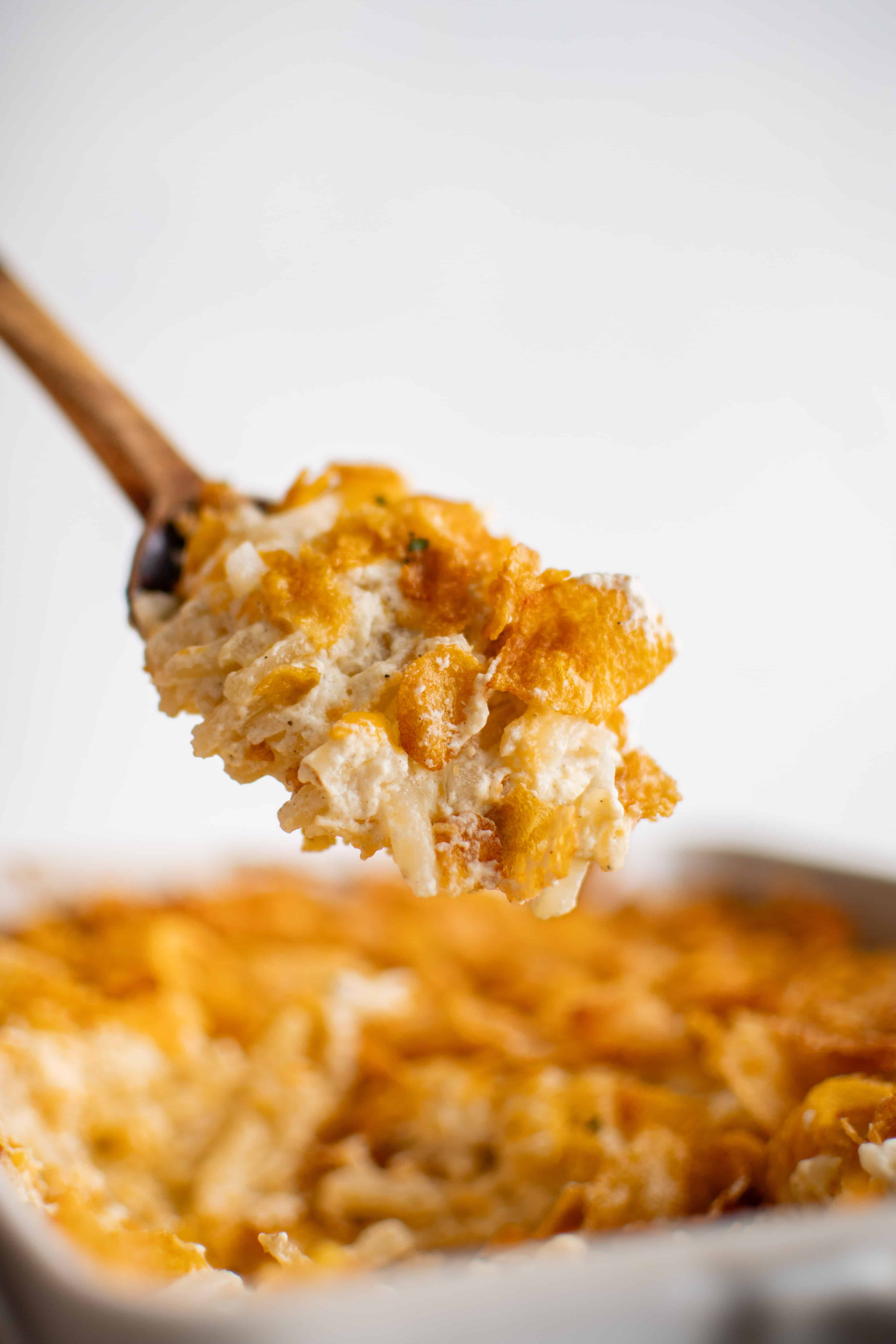 Wooden serving spoon filled with cheesy, creamy funeral potatoes hovering over a large casserole dish filled with the remaining baked funeral potatoes.