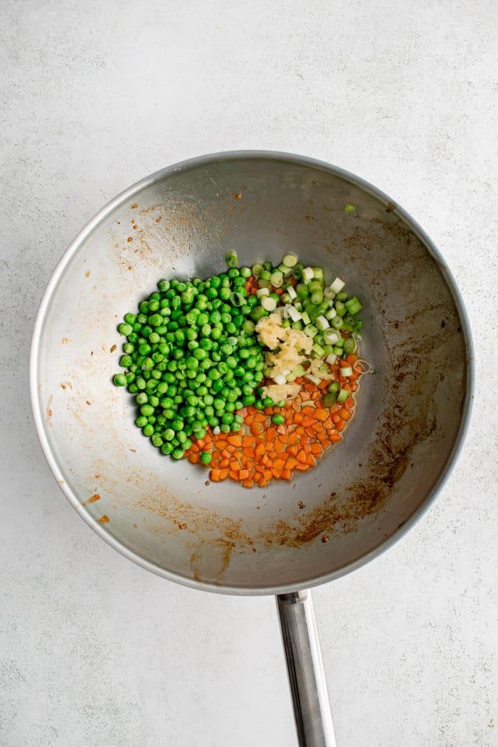 Large wok filled with diced carrots, frozen peas, chopped green onion, and minced garlic.