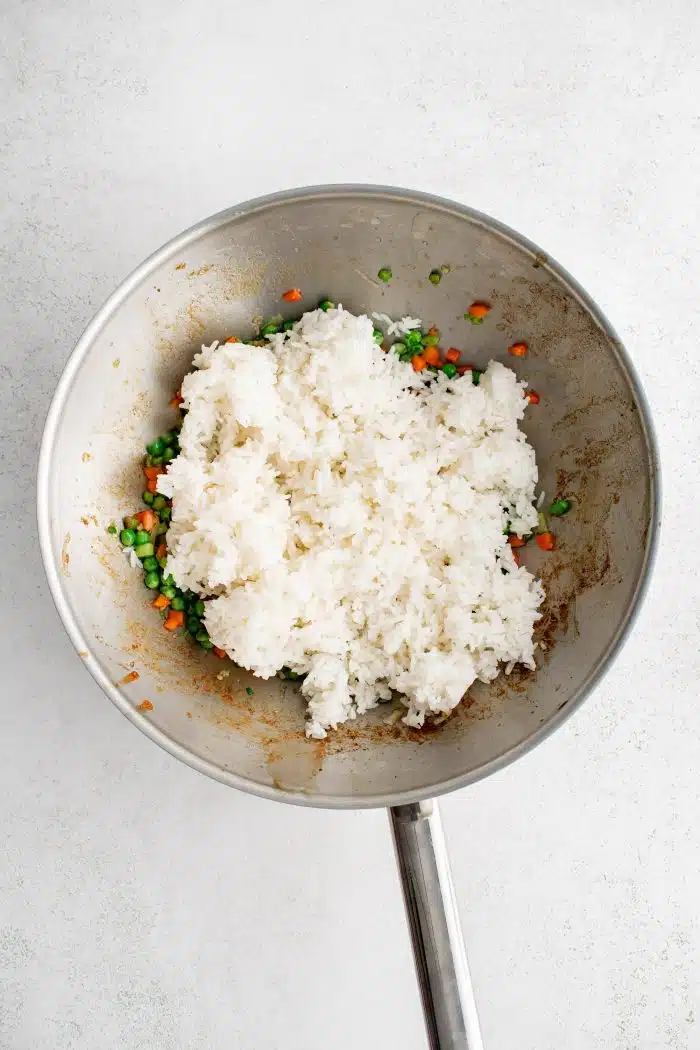 Cold, day-old white rice added to a large wok filled with cooked and softened diced carrots, frozen peas, garlic, and green onions.