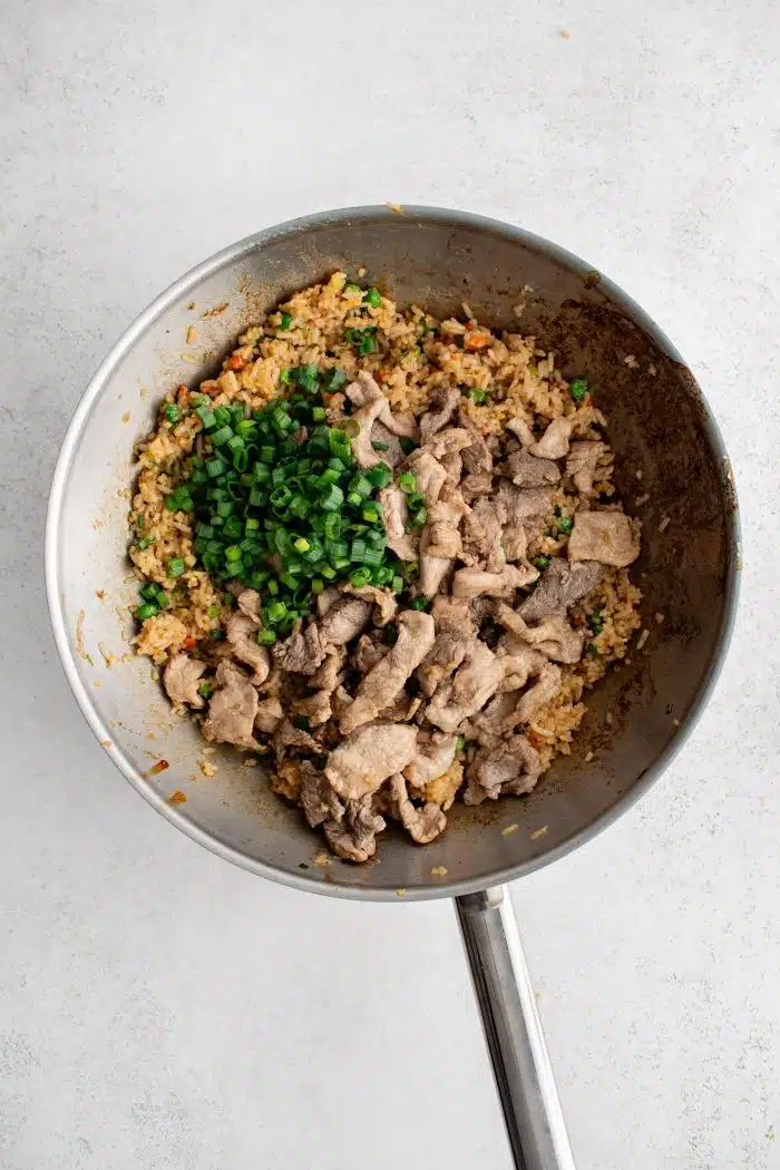 Cooked slices of marinated pork tenderloin and freshly chopped green onion added to a large wok filled with stir-fried rice.
