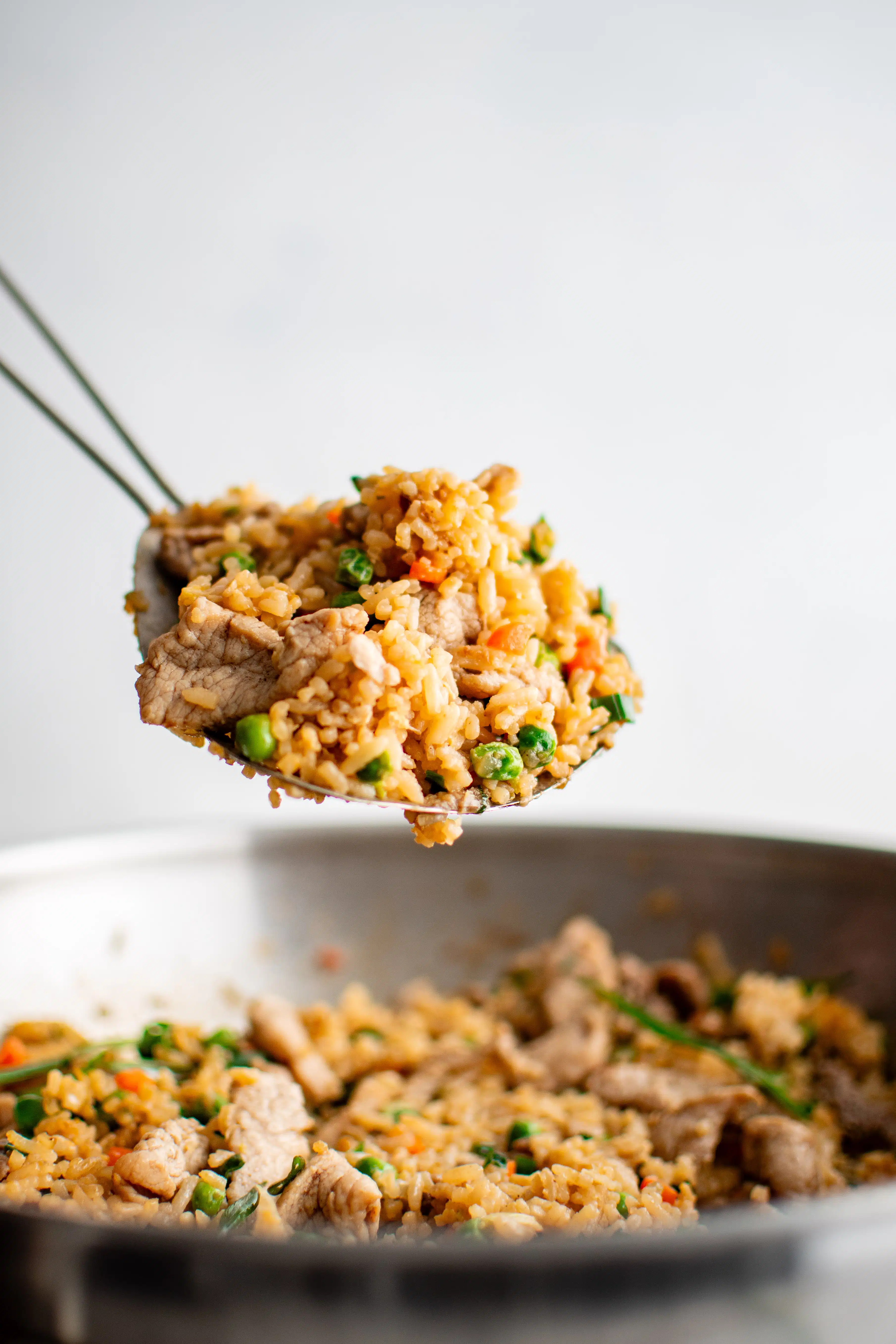 Large stainless steel spoon scooping pork fried rice from a large wok that is filled with cooked pork fried rice.