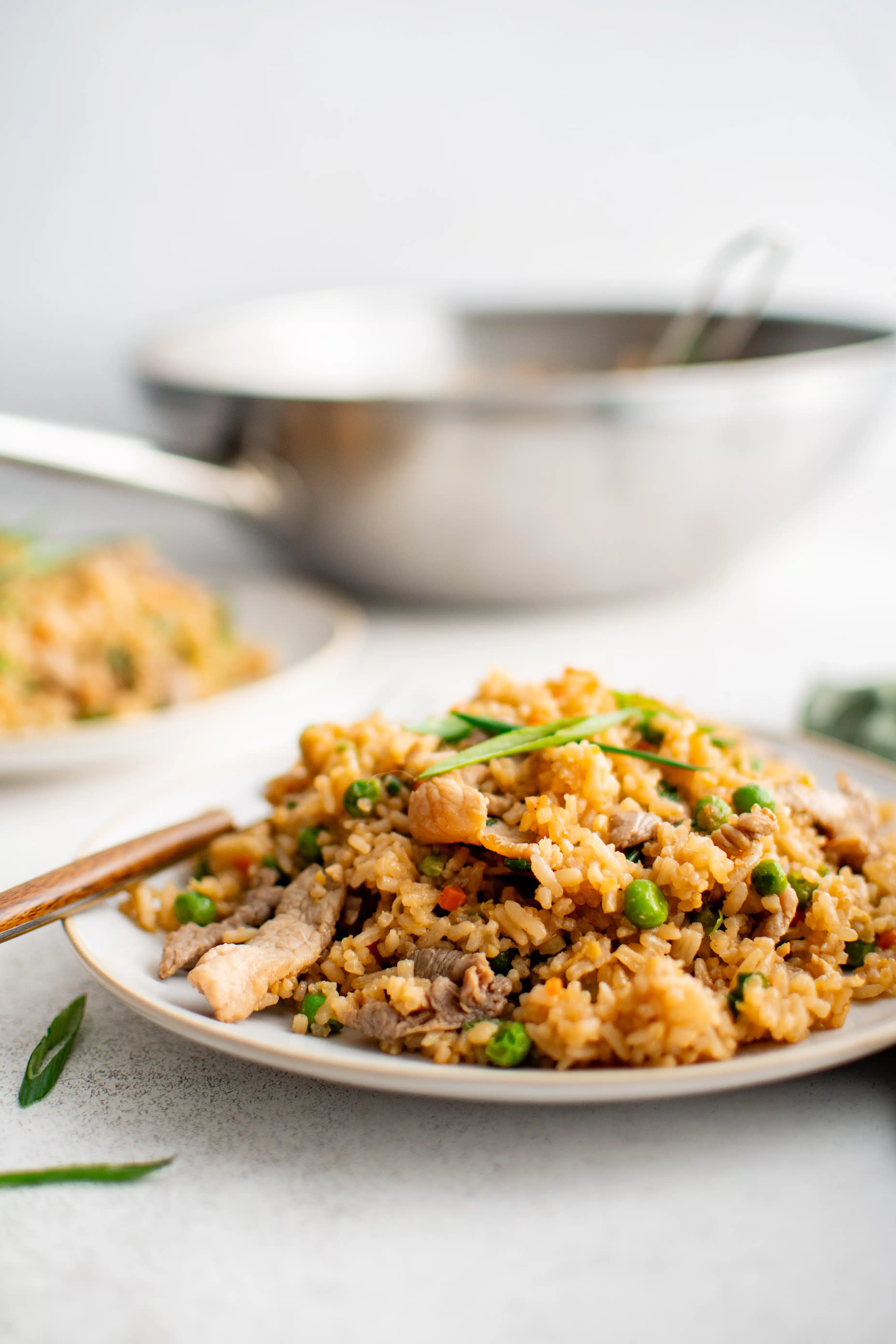 White serving plate with golden brown cooked pork fried rice garnished with green onion with a large stainless steel wok in the background.