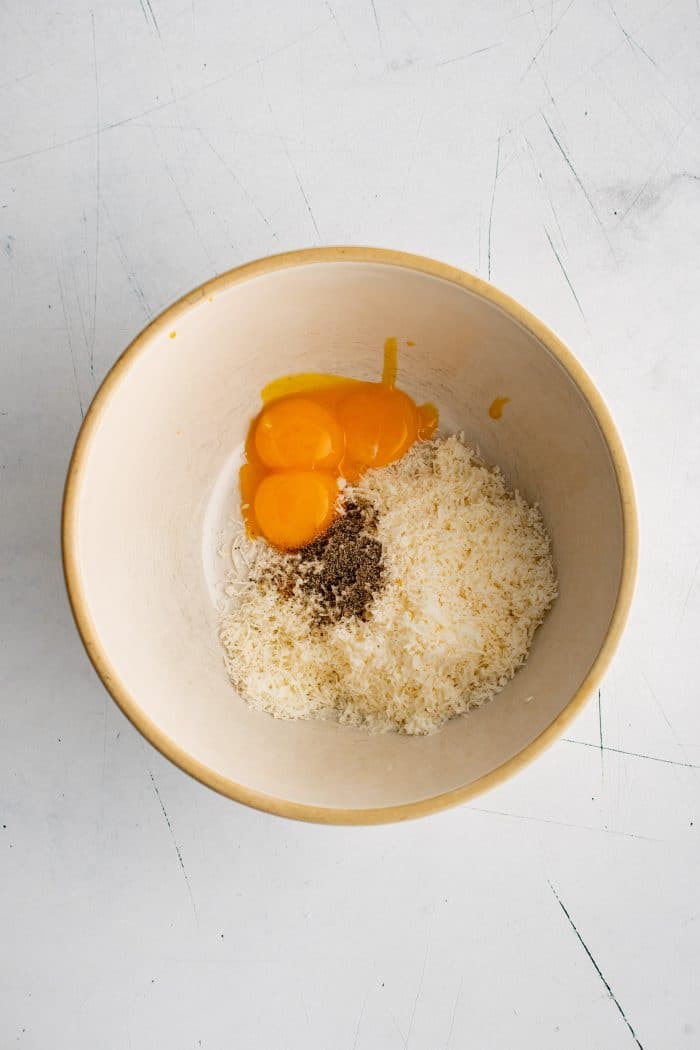 Three egg yolks, freshly grated Pecorino Romano cheese, and black pepper in a medium mixing bowl.