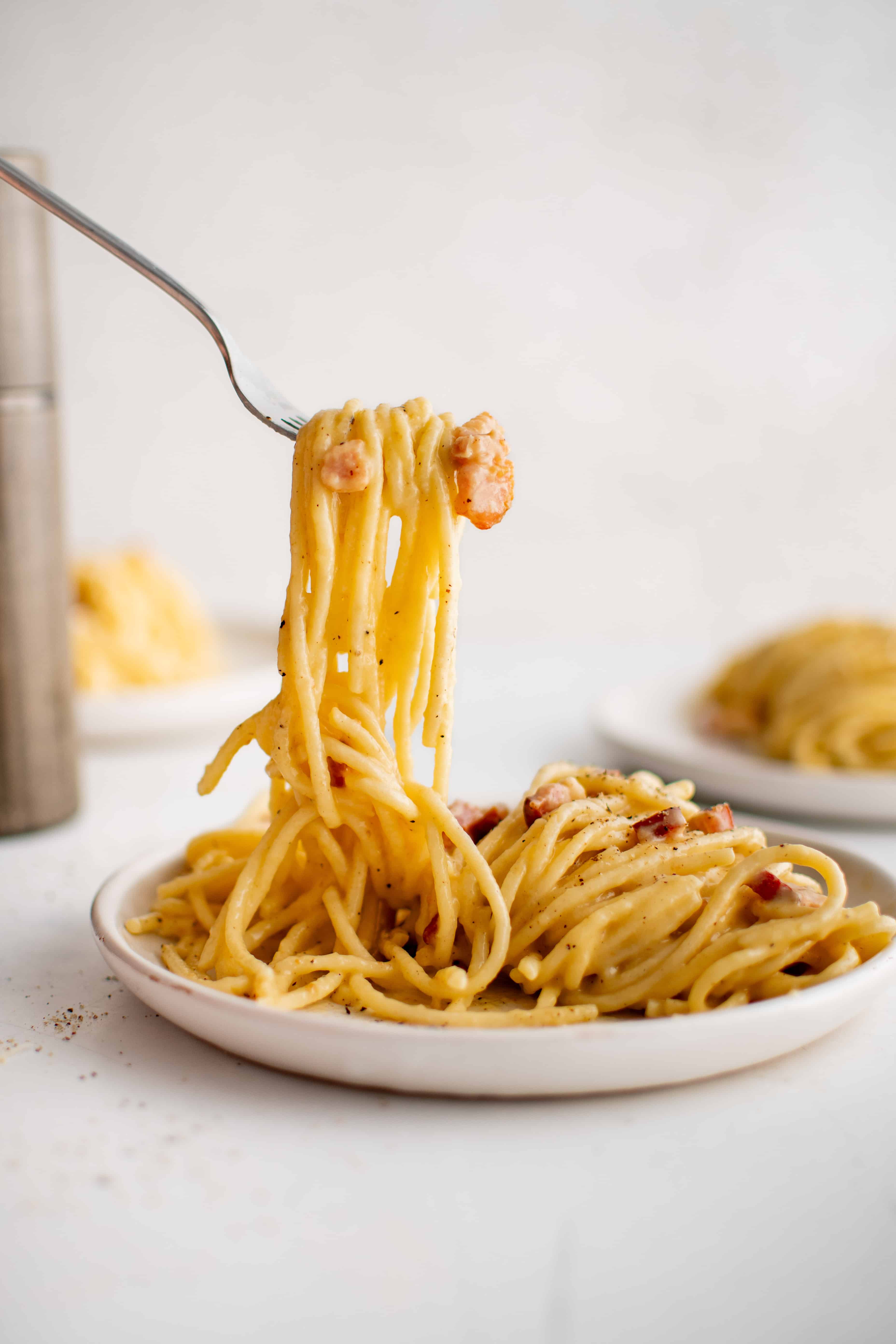 Fork filled with long strands of cooked spaghetti carbonara hovering over a plate filled with cooked pasta.
