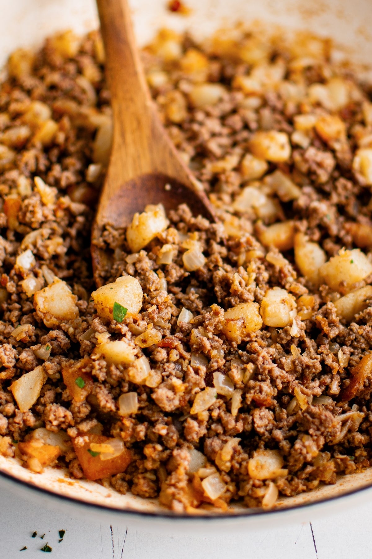 Close-up of ground beef, browned potato pieces, and diced onion in a cream-colored enameled skillet with a wooden spoon and small pieces of chopped green parsley.