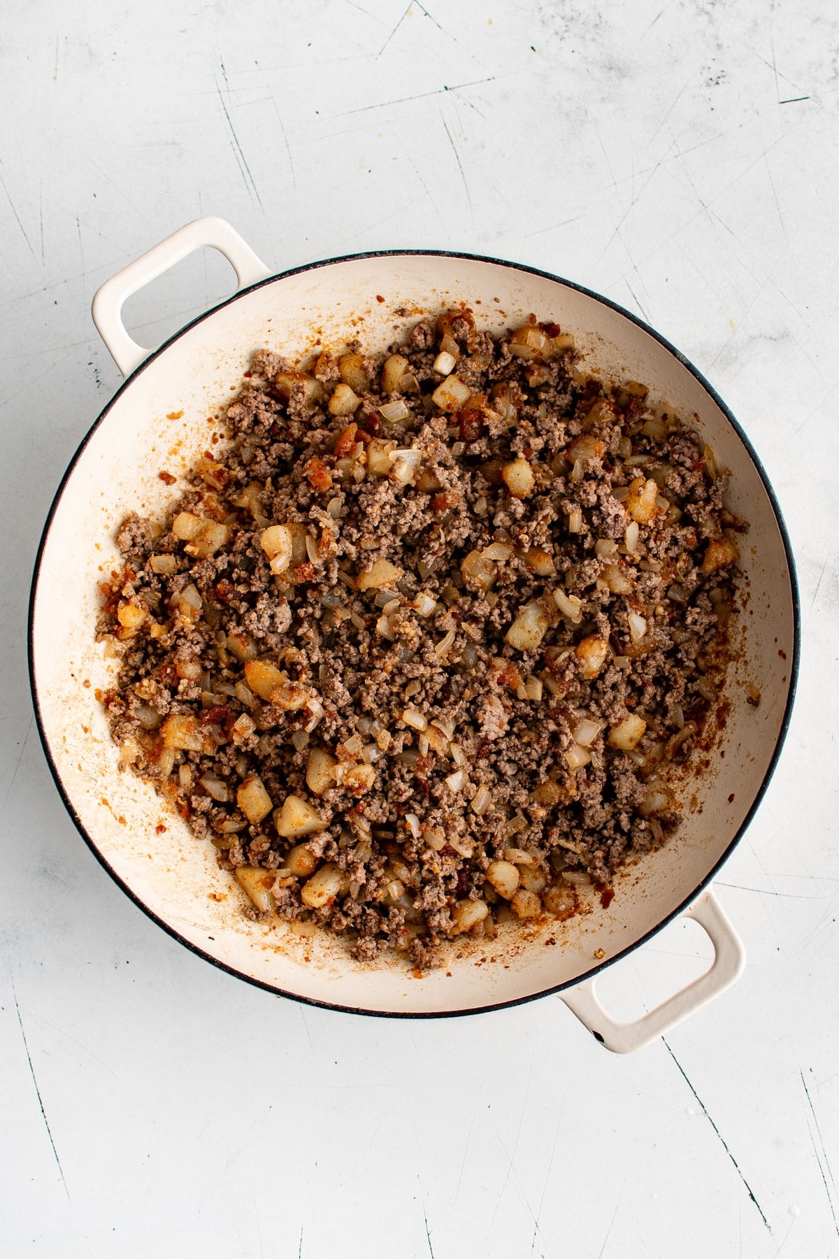 Overhead view of finished ground beef and golden potatoes mixed with diced onion in a large cream-colored enameled cast iron braiser.