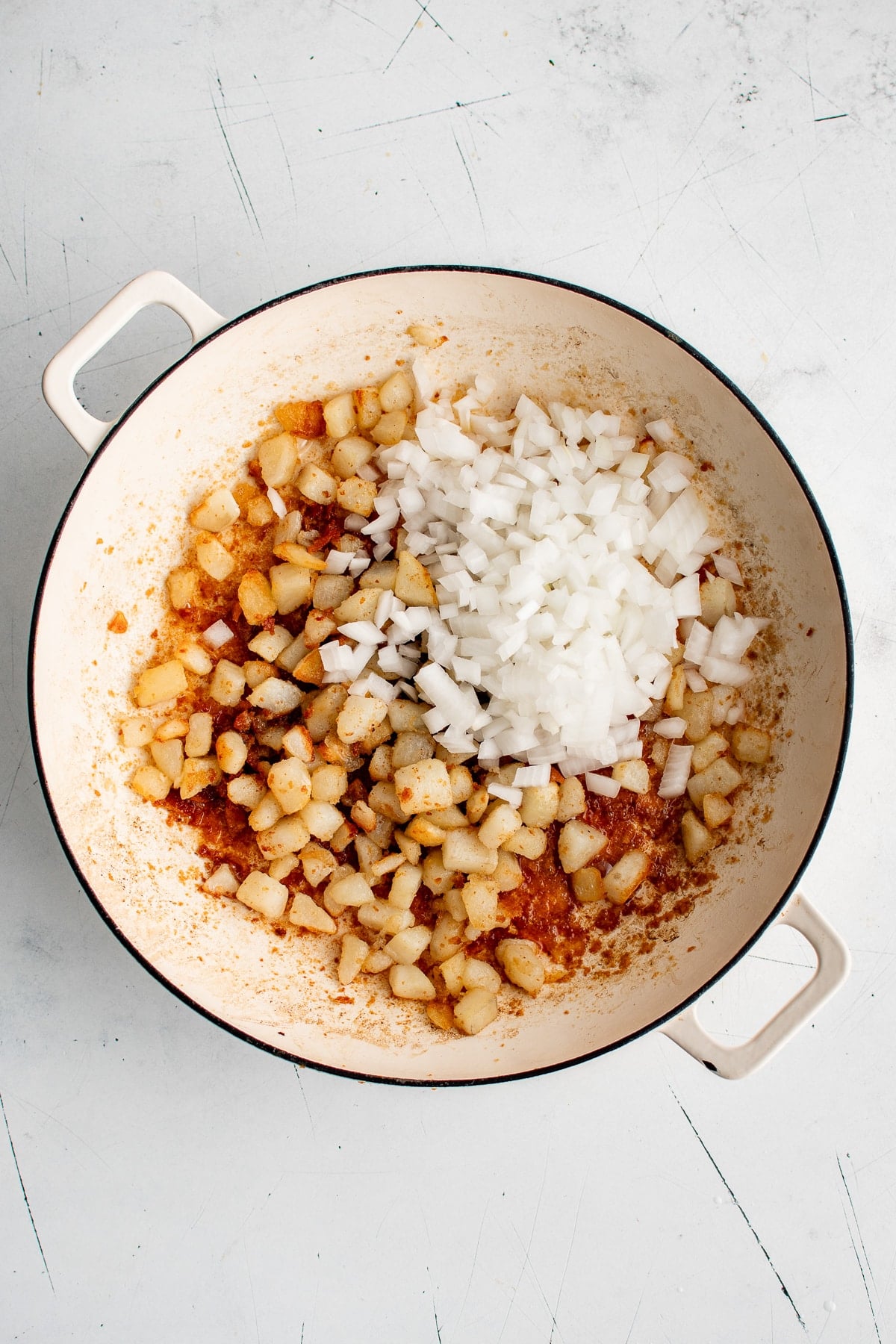 Overhead view of browned diced Yukon Gold potatoes in a cream-colored braiser with a mound of freshly diced white onion added on top.