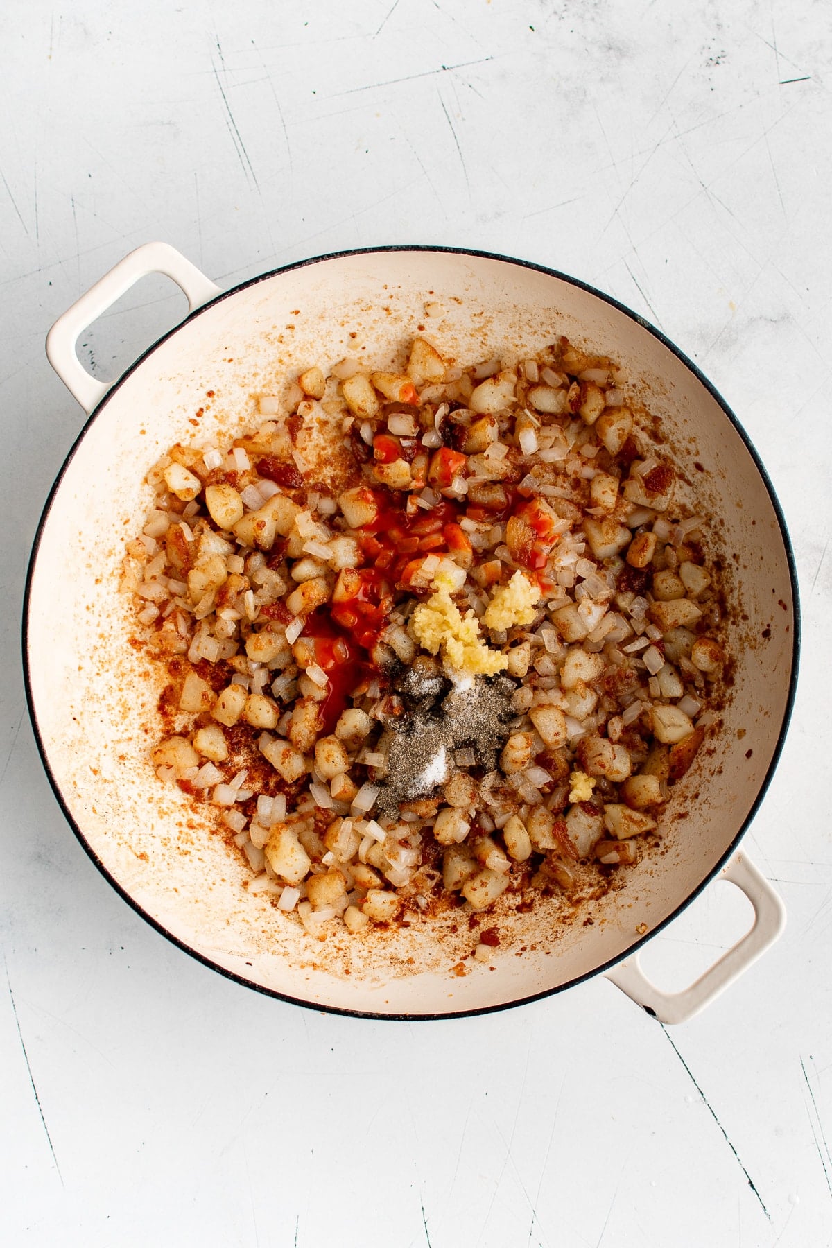 Overhead view of browned potatoes and softened onions in a cream-colored braiser topped with minced garlic, red hot sauce, salt, and black pepper.