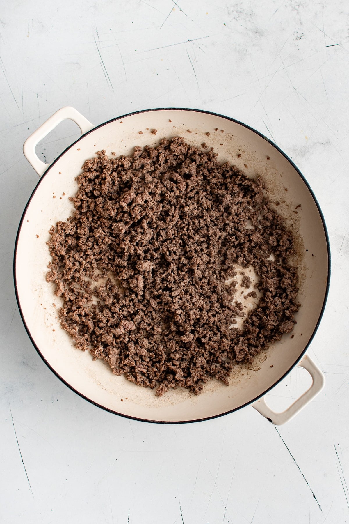 Overhead view of browned ground beef crumbled inside a large cream-colored enameled cast iron braiser with white handles.