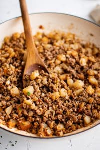 Close angled view of seasoned ground beef, golden potato pieces, and diced onions in a cream-colored skillet with a wooden spoon and a light parsley garnish.