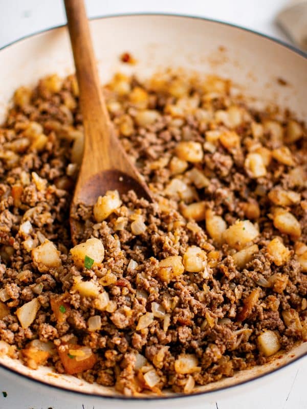 Close angled view of seasoned ground beef, golden potato pieces, and diced onions in a cream-colored skillet with a wooden spoon and a light parsley garnish.