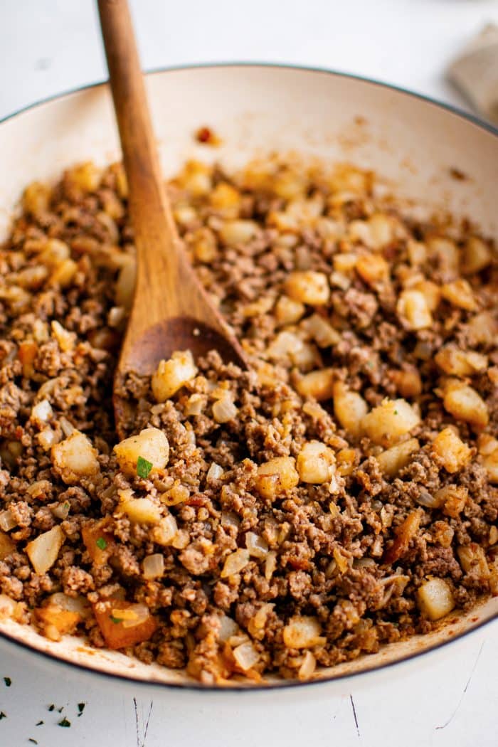 Close angled view of seasoned ground beef, golden potato pieces, and diced onions in a cream-colored skillet with a wooden spoon and a light parsley garnish.