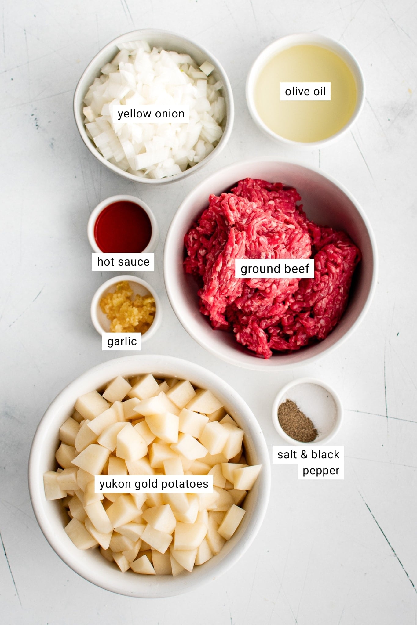 Overhead ingredient photo showing raw ground beef, peeled diced Yukon Gold potatoes, diced yellow onion, olive oil, red hot sauce, minced garlic, salt, and black pepper in white bowls. White text labels identify yellow onion, olive oil, hot sauce, ground beef, garlic, Yukon Gold potatoes, and salt and black pepper.