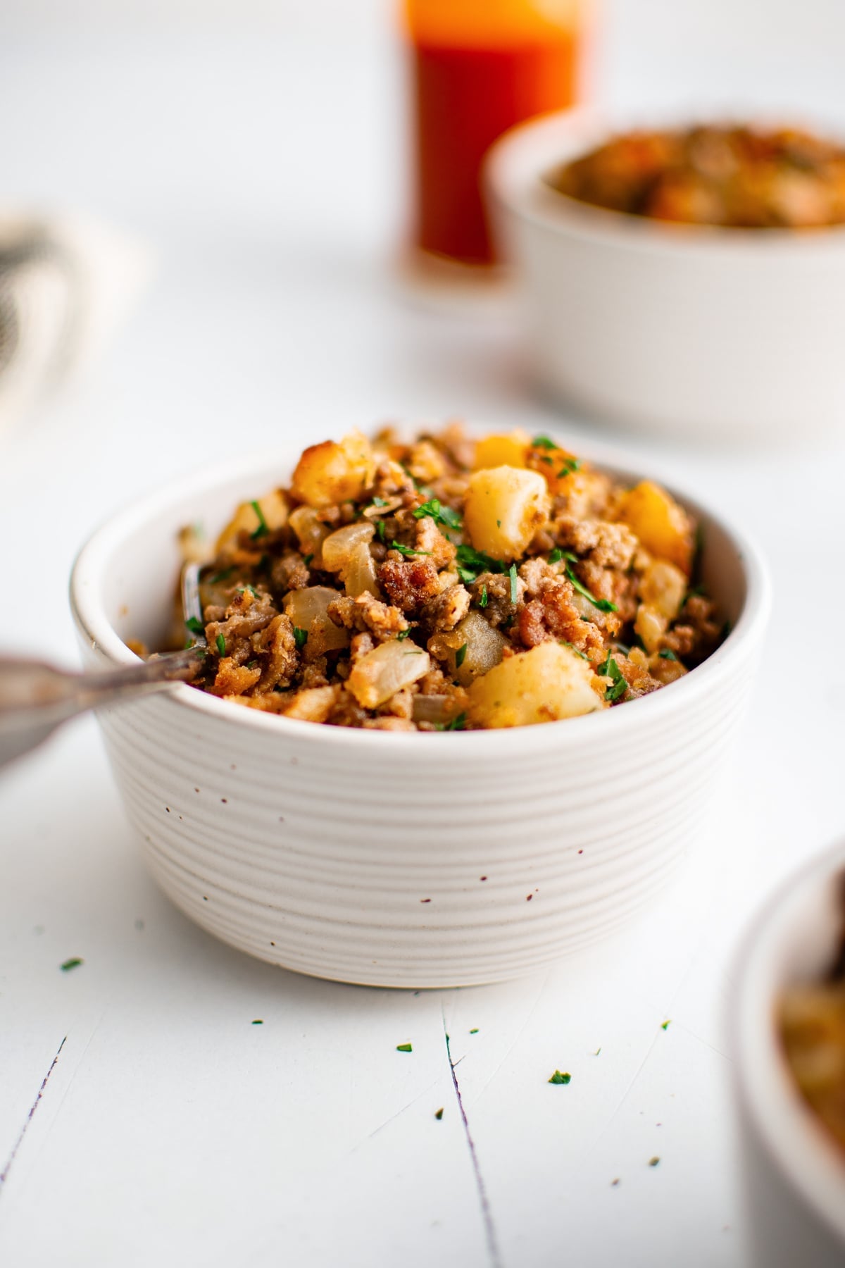 Ground beef and potatoes served in a white ribbed ceramic bowl and garnished with chopped green parsley, with a spoon in the bowl, a hot sauce bottle blurred in the background, and additional white serving bowls behind it.
