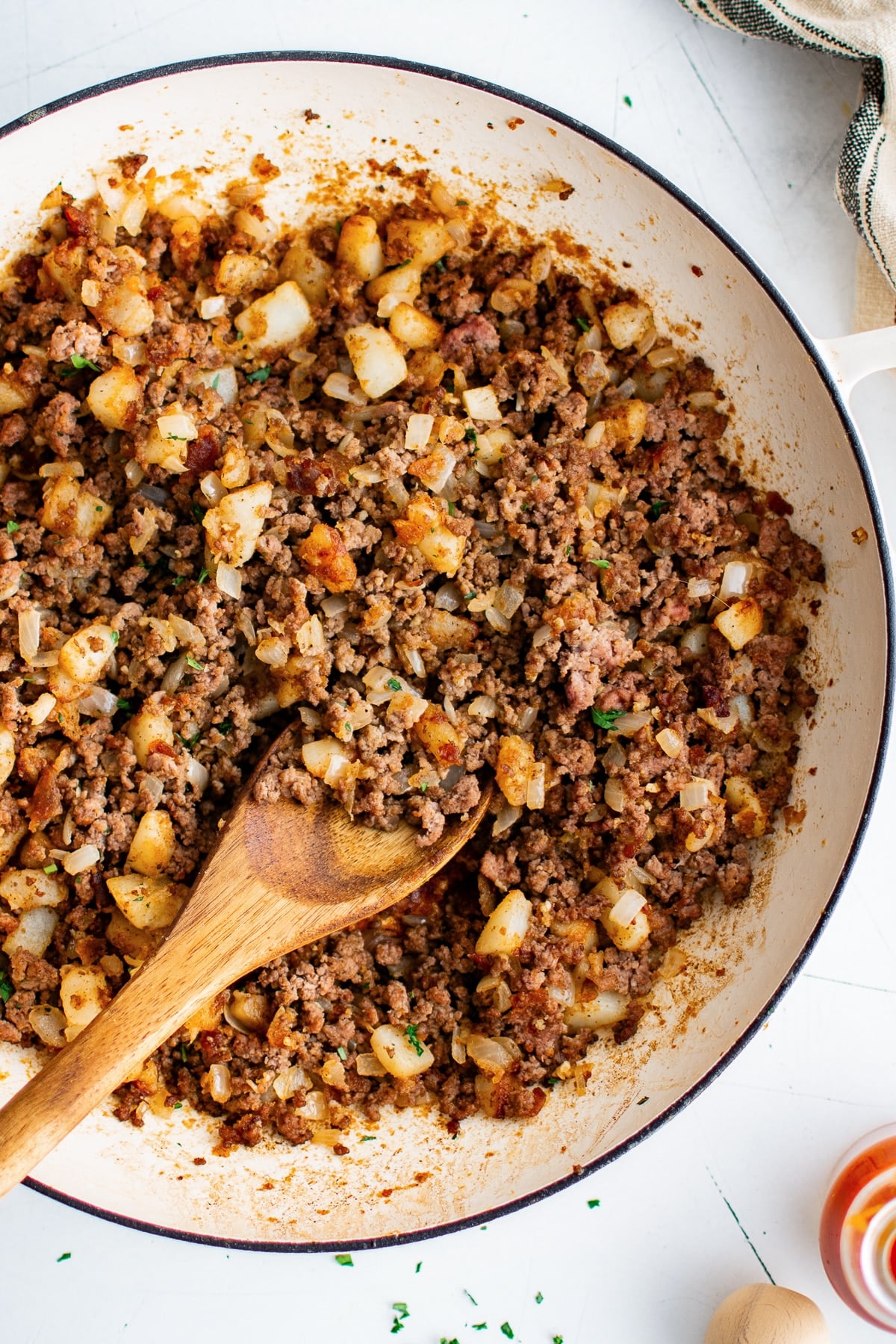 Close overhead view of ground beef and golden potatoes in a cream-colored braiser with a wooden spoon resting in the skillet and chopped green parsley sprinkled over the dish.