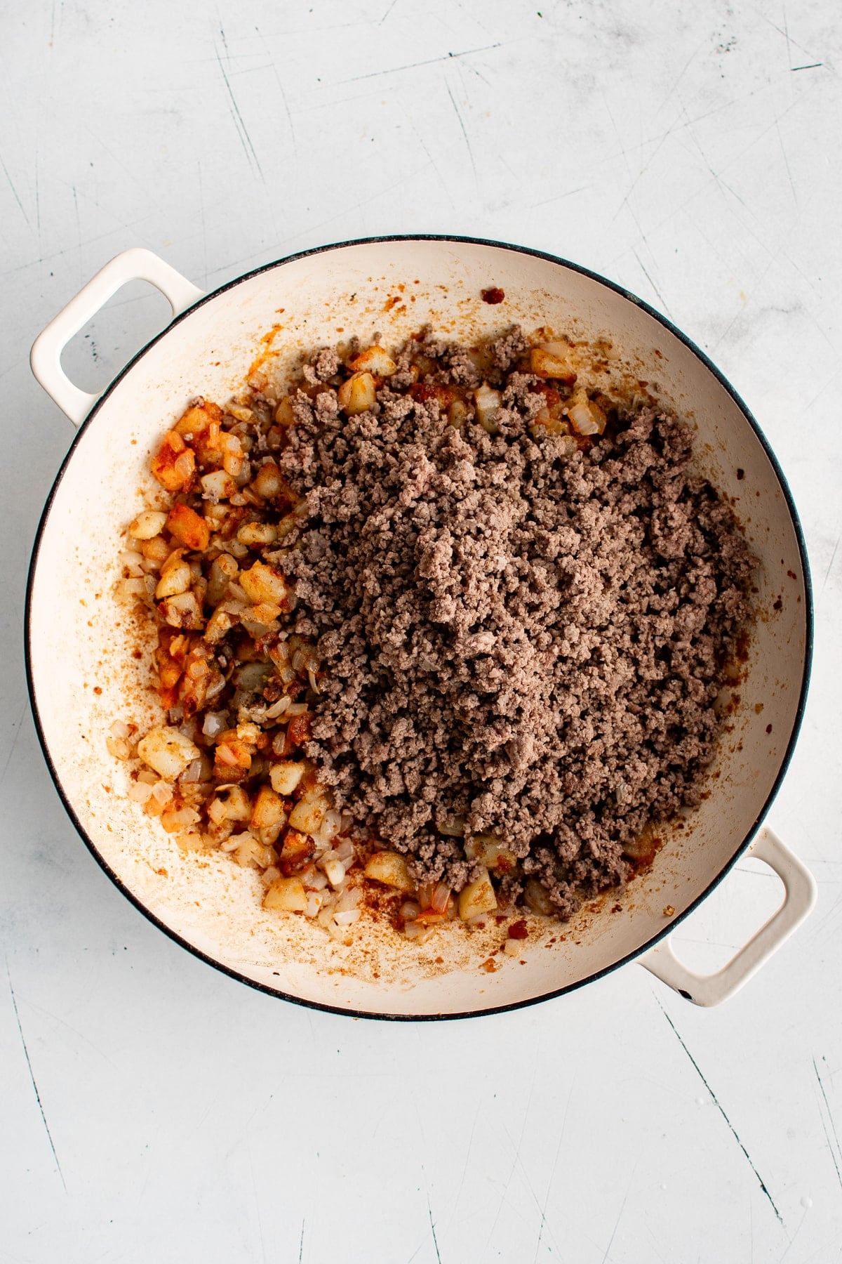 Overhead view of cooked ground beef being returned to browned potatoes and onions in a cream-colored enameled cast iron braiser.