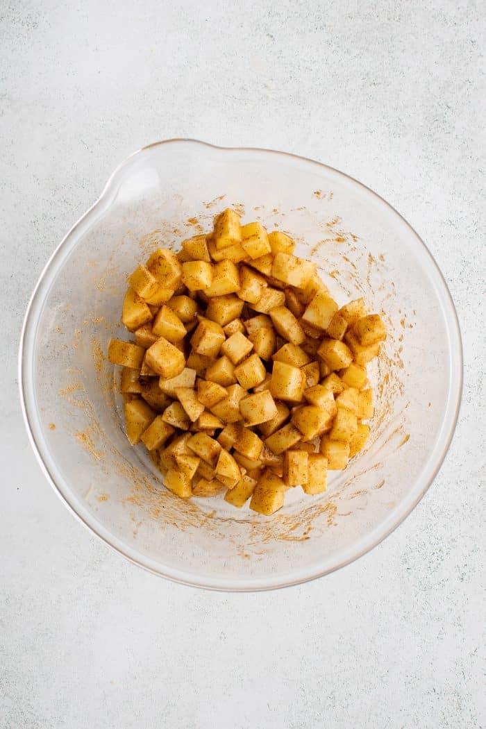 A large glass mixing bowl filled with raw diced and peeled Yukon gold potatoes coated in oil and spices.