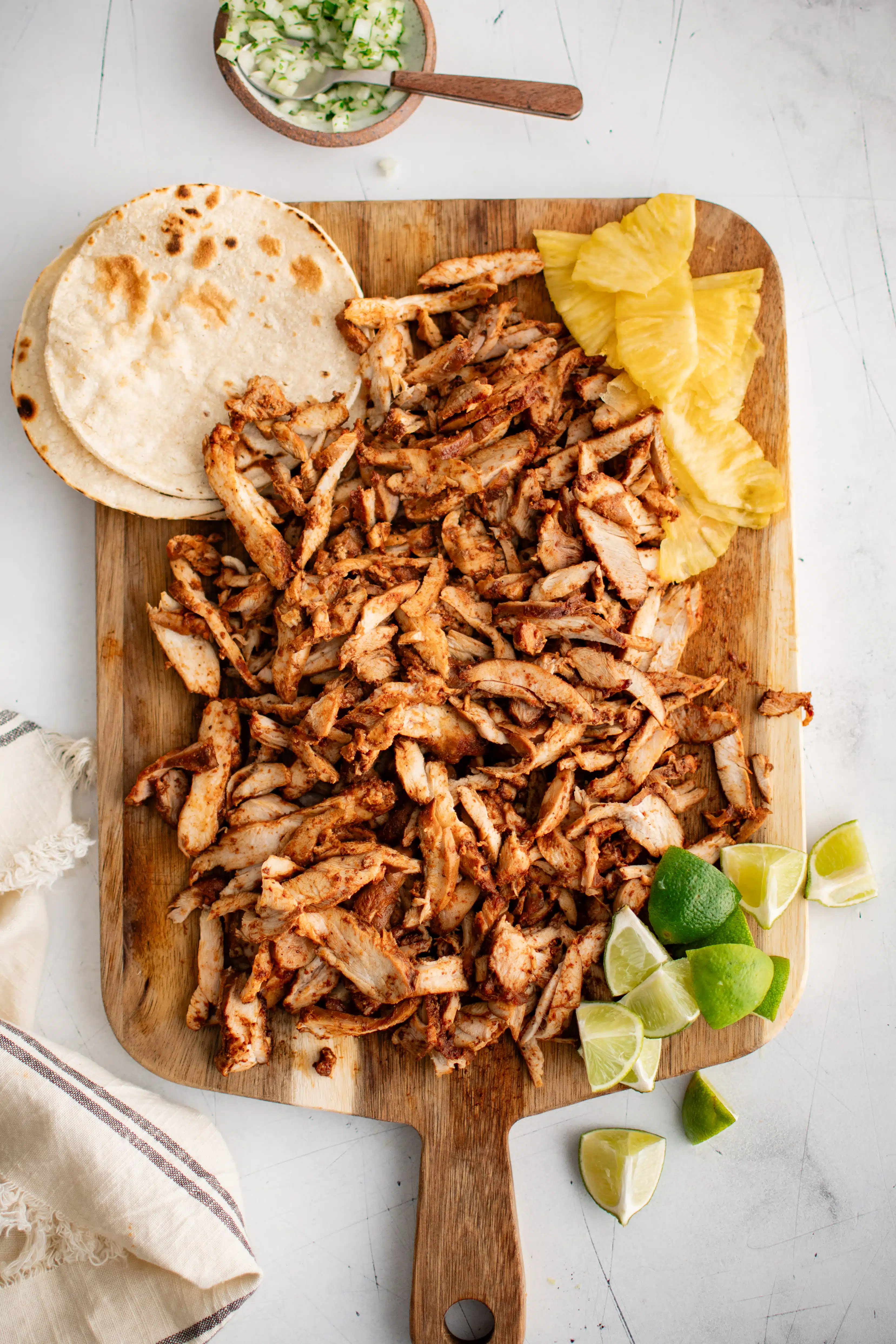 Thinly sliced chicken al pastor arranged over a large cutting board and served with sliced pineapple, flour tortillas, and lime wedges.