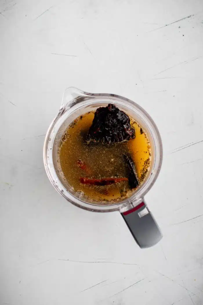 Chicken broth, pineapple juice, orange juice, lime juice, and reconstituted arbol chiles and guajillo chiles added to the blender filled with whole garlic cloves, roughly chopped white onion, Mexican oregano, salt, thyme, achiote paste, cumin powder, ground black pepper, and ground cloves.