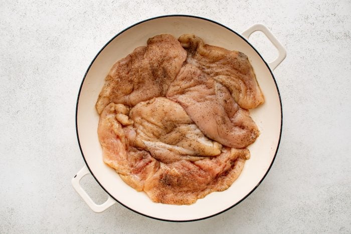 Large white oven-safe pan filled with raw, uncooked chicken breasts seasoned with salt and black pepper.