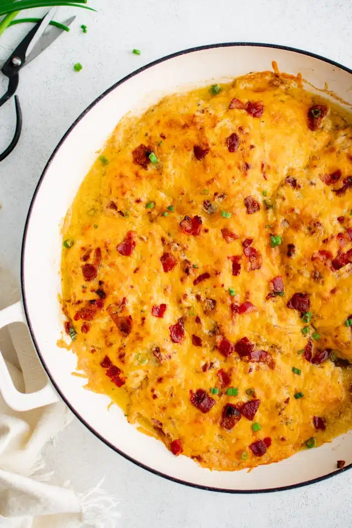 Large white oven-safe pan filled with chicken breasts covered in a mixture of cream of chicken soup, cream cheese, ranch seasoning mix, gooey melted cheddar cheese, and bacon bits.