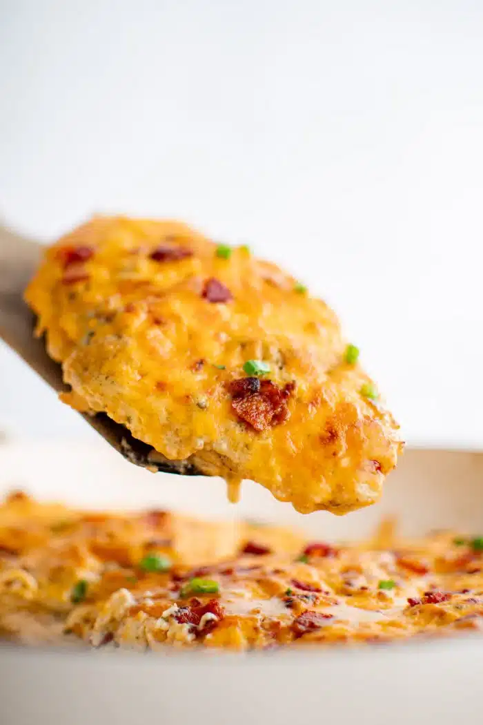 Wooden serving spatula removing a baked chicken breast covered with "crack" chicken combination of cream of bacon, chicken soup, cheddar cheese, ranch seasoning mix, and cream cheese.