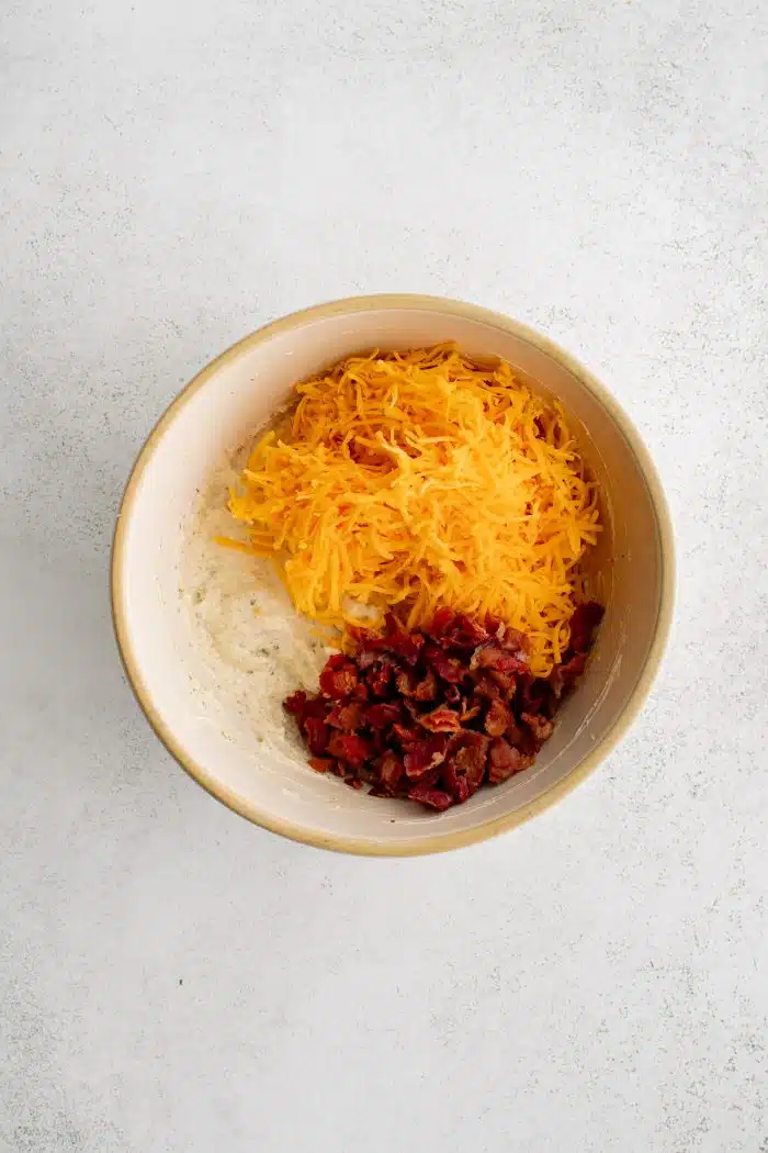 Shredded cheddar cheese and bacon bits added to a mixing bowl filled with the mixture of cream cheese, cream of chicken soup, and ranch seasoning mix.