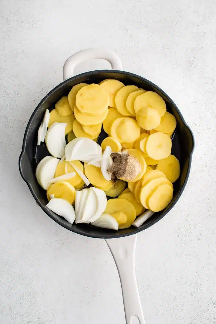 Large cast iron pan filled with raw peeled and sliced Idaho potatoes, vegetable oil, sliced onion, garlic powder, salt, and black pepper.